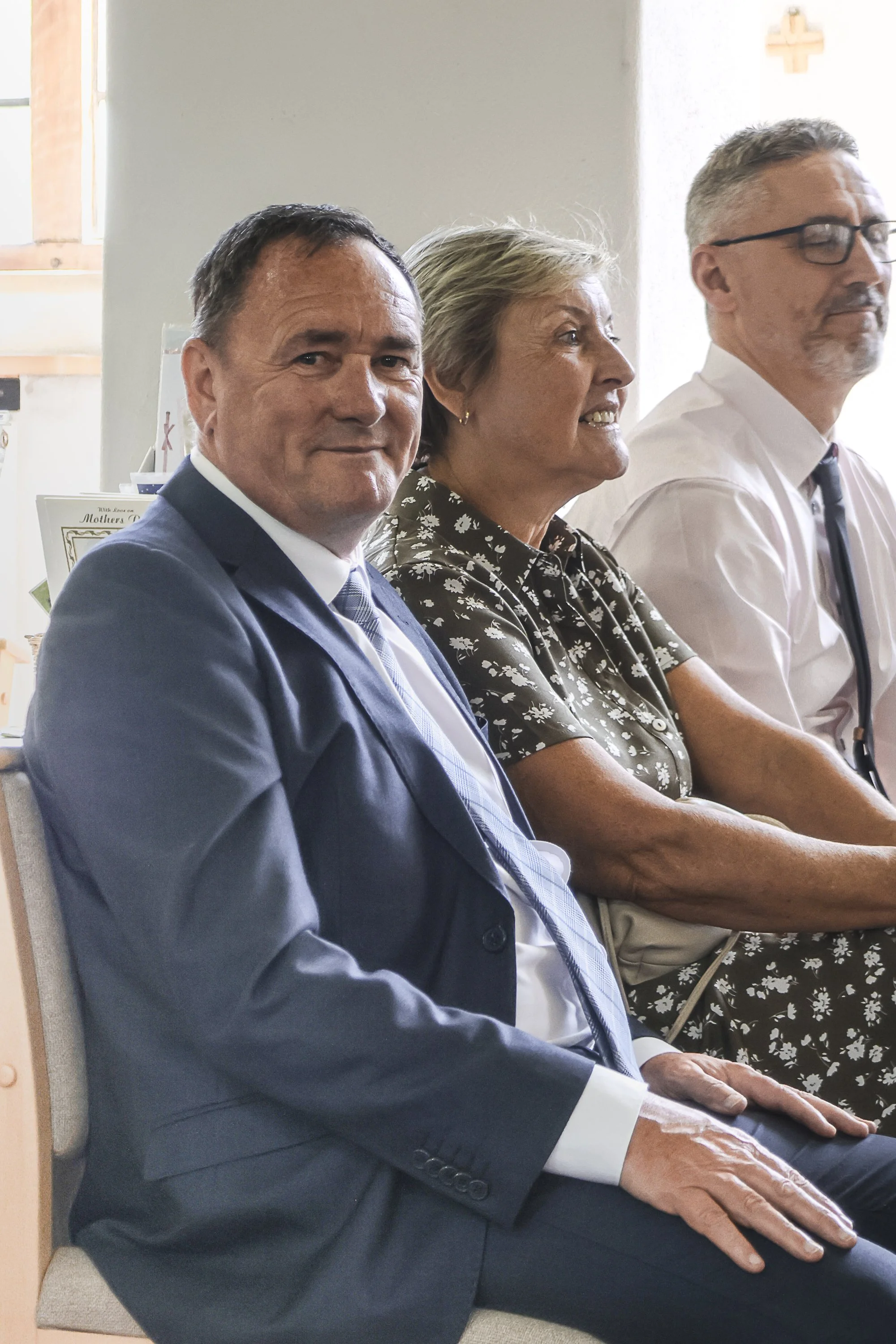 Three adults sitting in a row. The man on the left, in a suit, smiling at the camera. The woman in the middle, in a floral shirt, also smiling. The man on the right, wearing glasses and a white shirt, appears to be looking away.