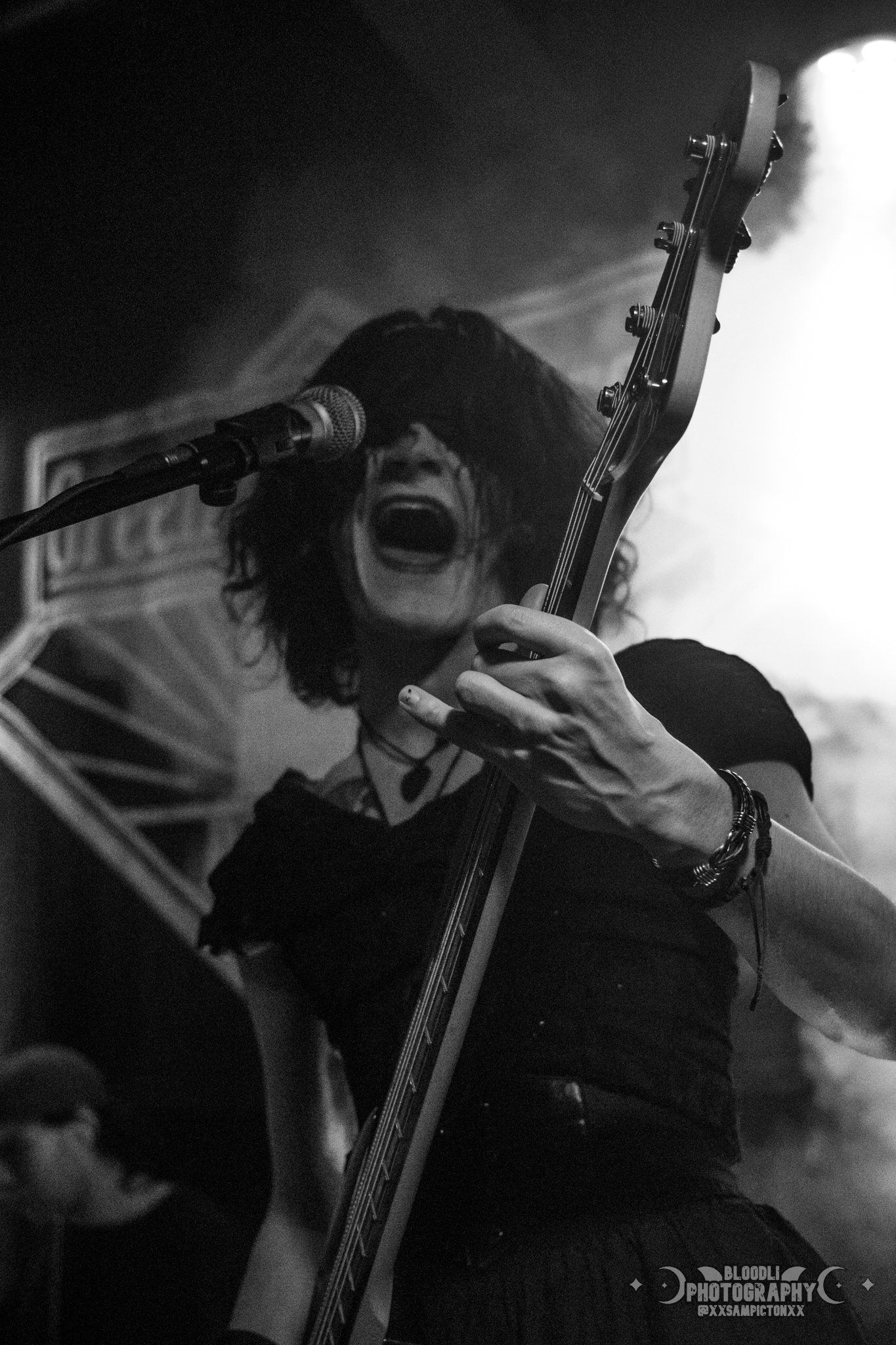 Black and white photo of a female musician passionately singing and playing an electric guitar on stage, with a microphone in front of her.