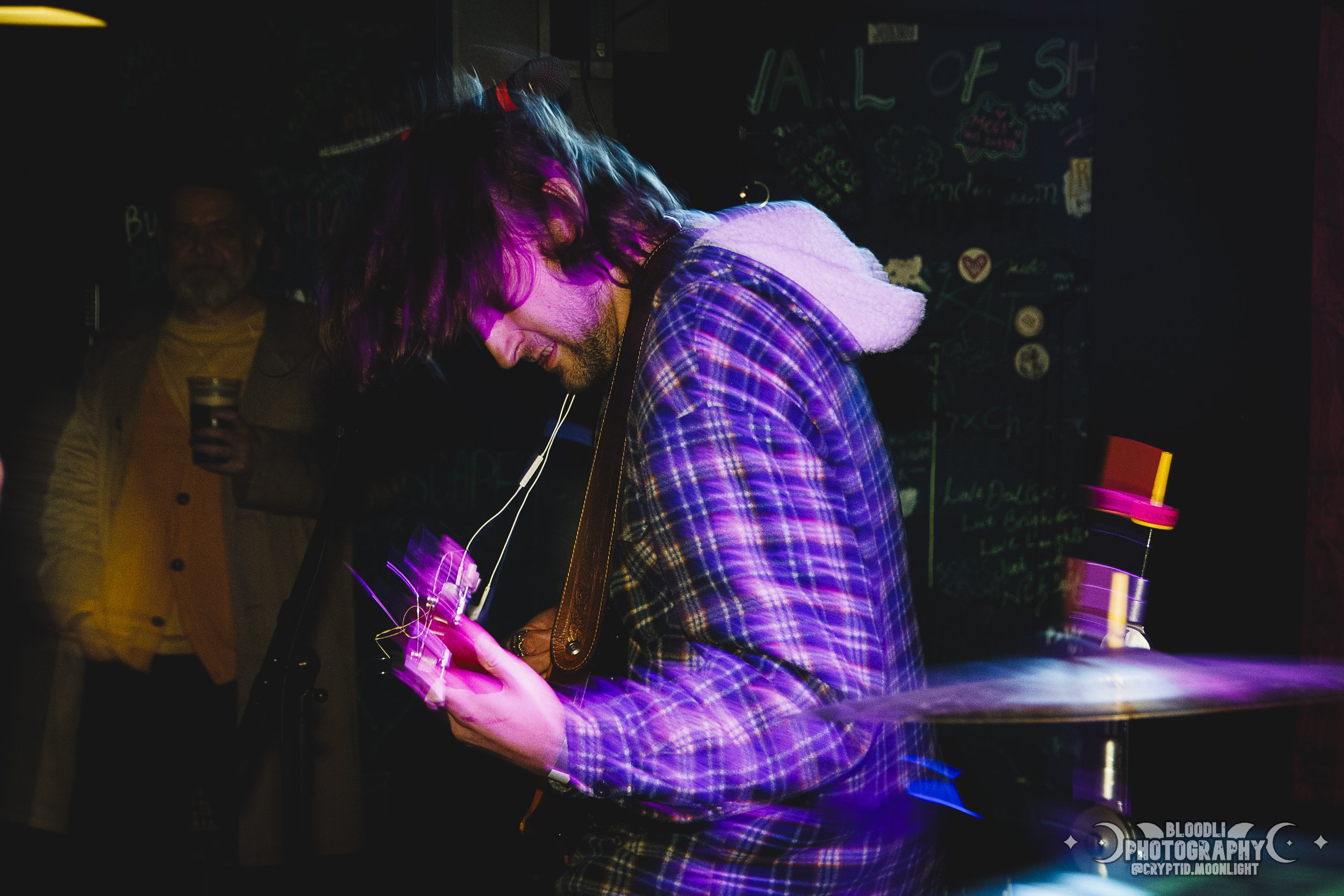 A man with long hair and a beard playing an electric guitar at a dark, colorful music venue.