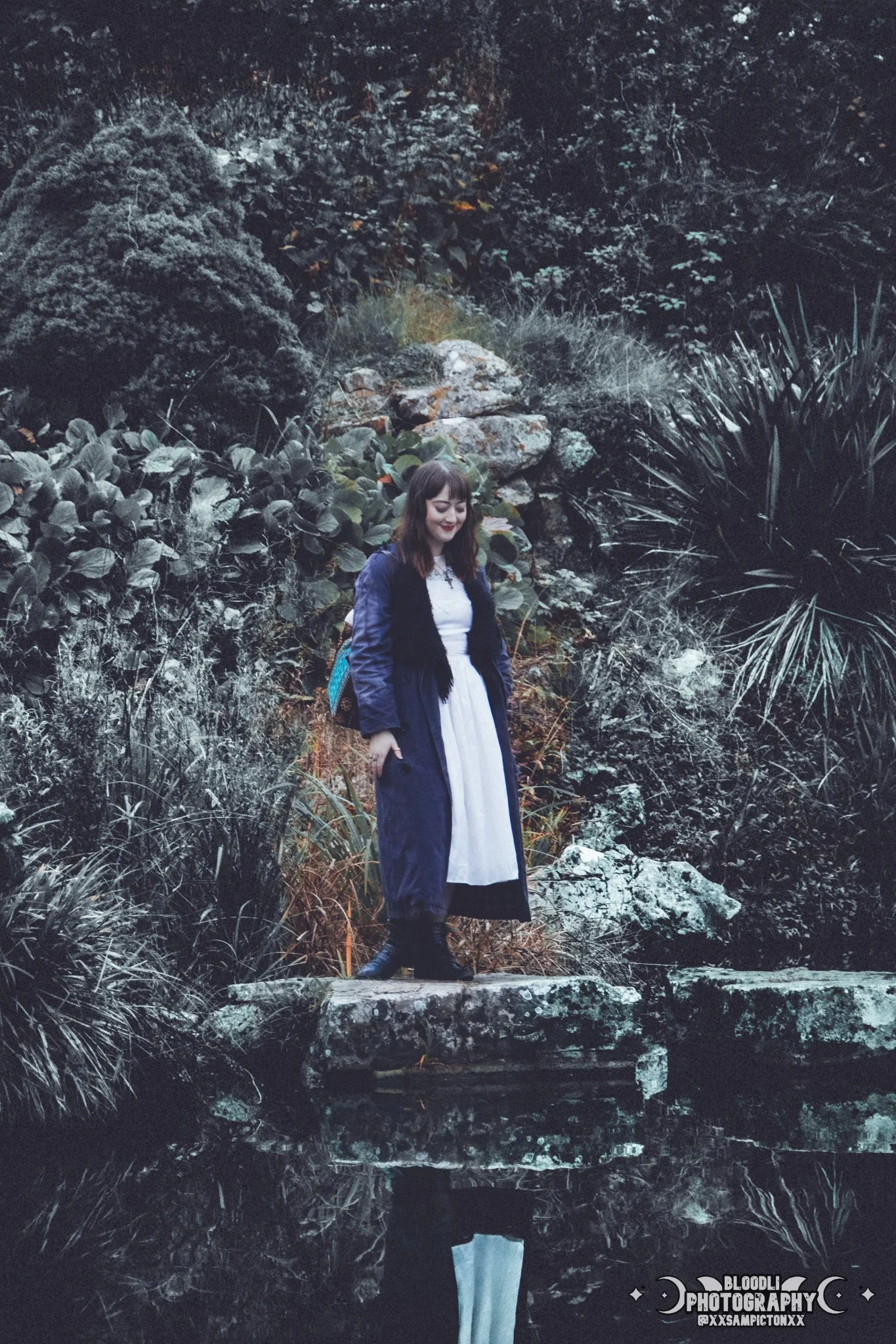 A young woman wearing a long white dress and a dark jacket with a fur collar, standing on a rock near a body of water, surrounded by dark green foliage and rocks, with her reflection visible in the water.