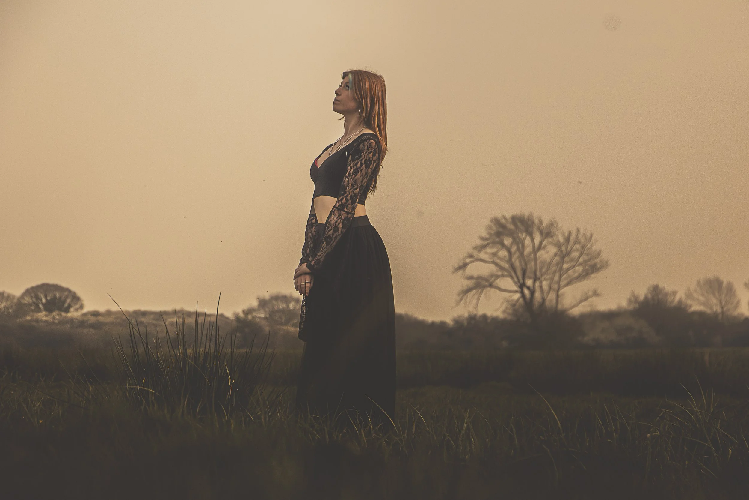 A woman wearing a black lace top and long black skirt standing in a field during sunset or dusk, with trees in the background.