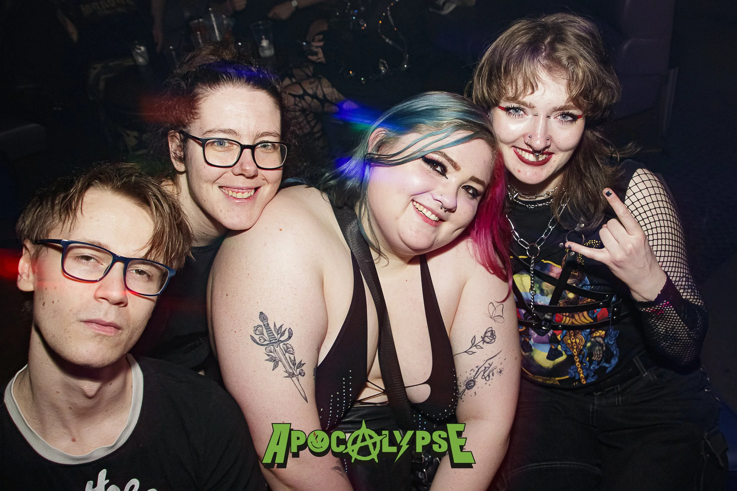 Group of five young adults at a gothic-themed event, with three women and two men posing together indoors, some displaying tattoos and gothic fashion.