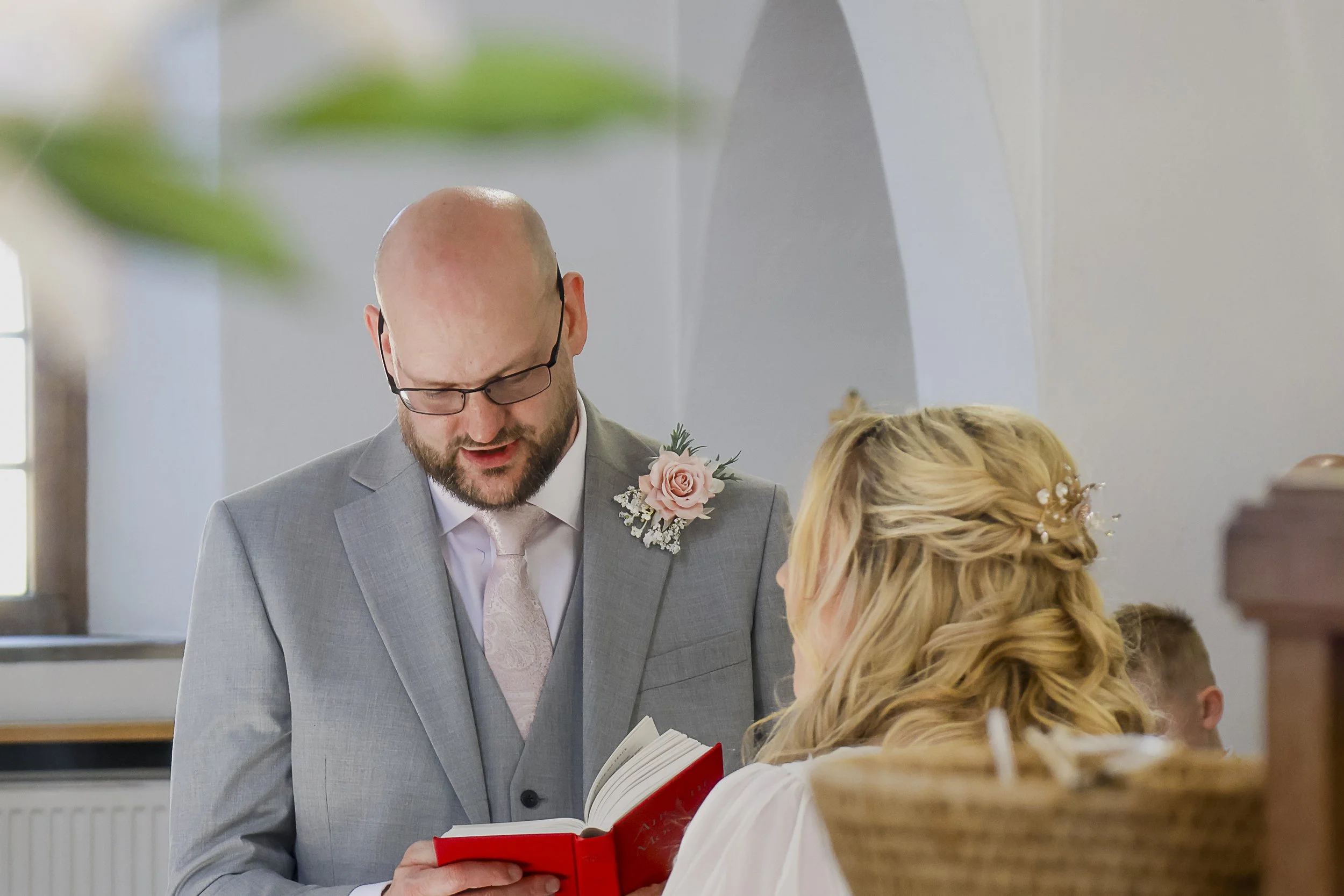 A man in a gray suit with a pink tie and glasses is holding a small red book and looking down, while a woman with blonde curly hair, wearing a white dress, is signing a document. They appear to be at a wedding ceremony.