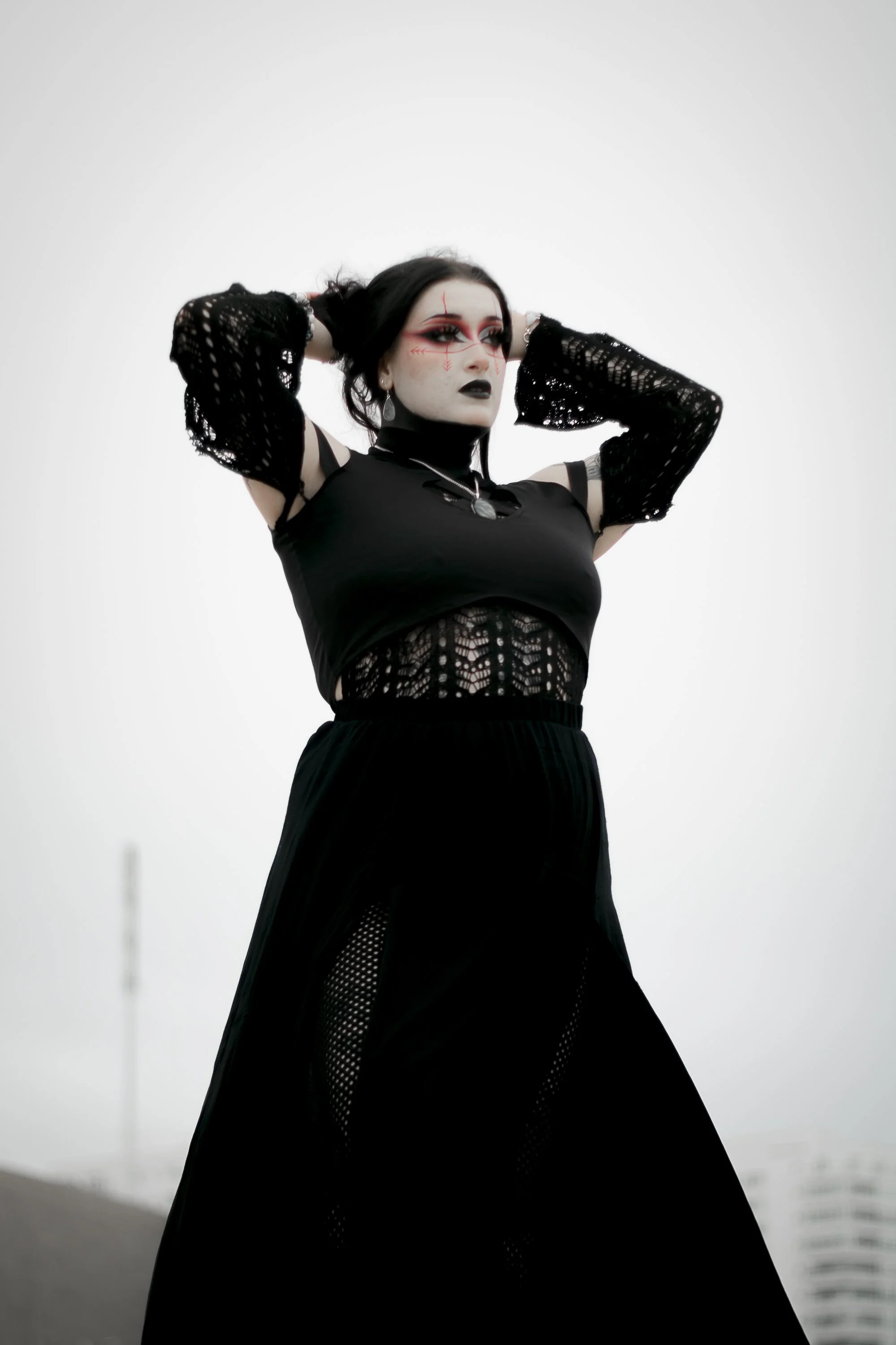 A woman with gothic makeup and black hair, wearing a black lace top and skirt, posing with her hands in her hair against a plain, overcast sky background.