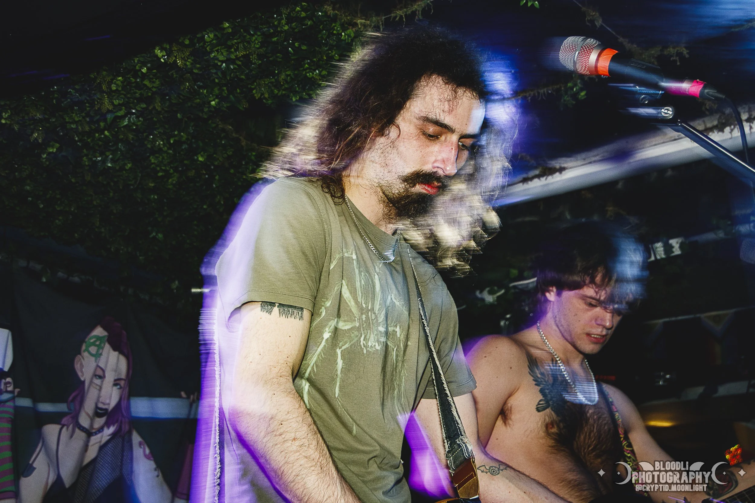 Two musicians perform at a nightclub, one with long hair and a beard playing guitar, and the other shirtless with tattoos. Both are under colorful lighting and appear focused on their instruments.