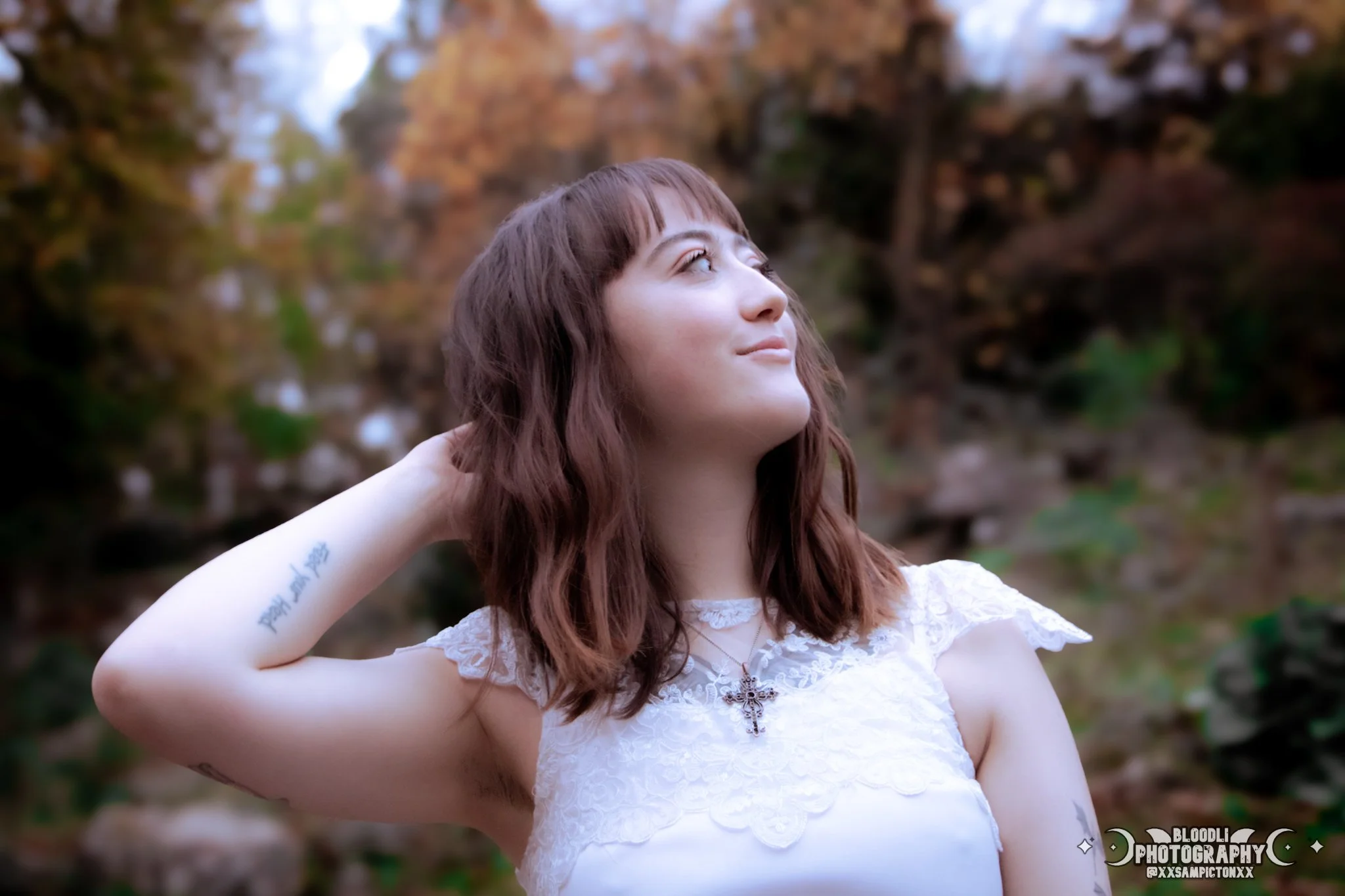 A young woman with wavy brown hair and tattoos on her arm, wearing a white lace dress with a cross necklace, standing outdoors with autumn-colored trees in the background, looking up and smiling.