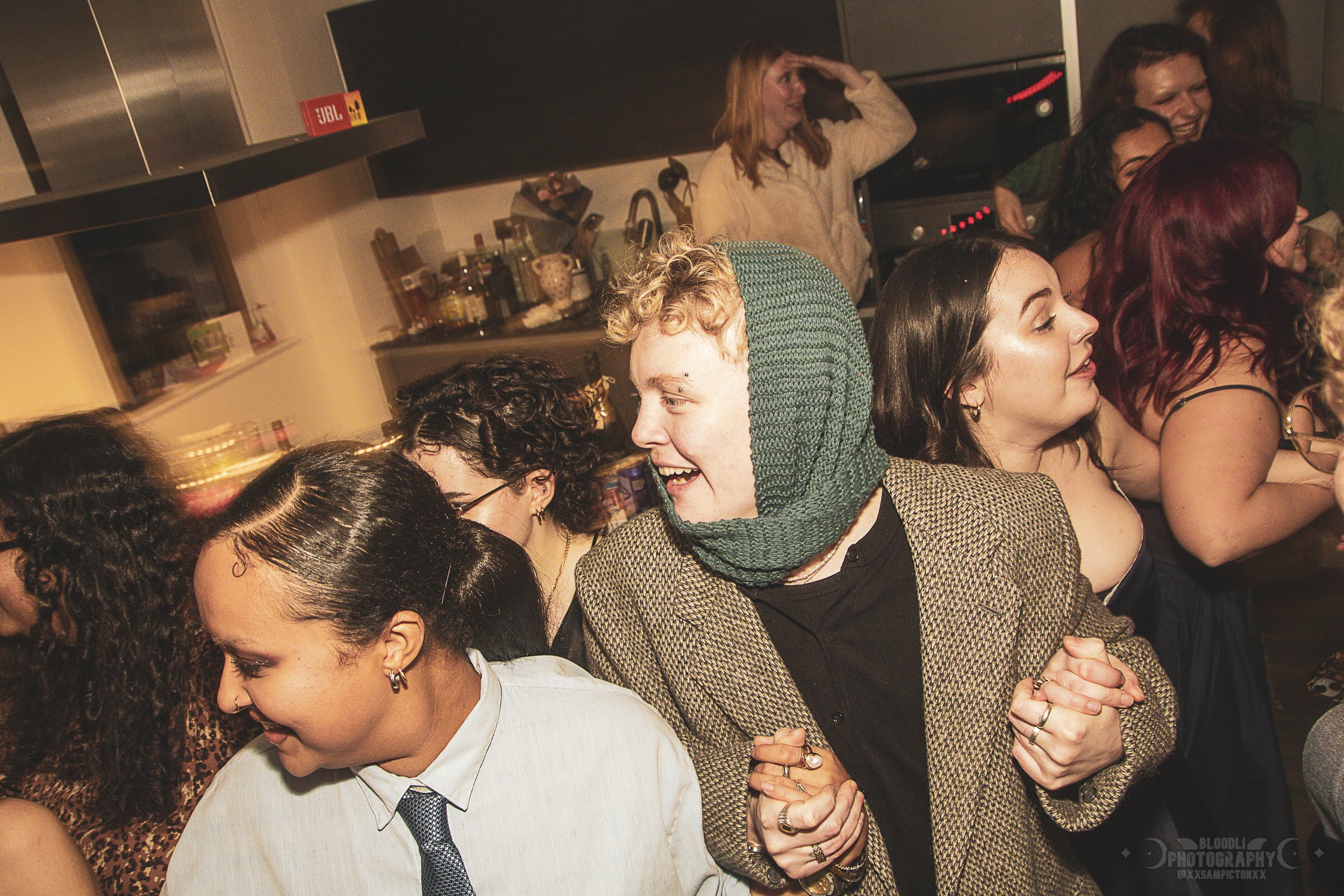 A group of people at a party or gathering, smiling and holding hands, in a kitchen setting with a person saluting or shielding their eyes in the background.