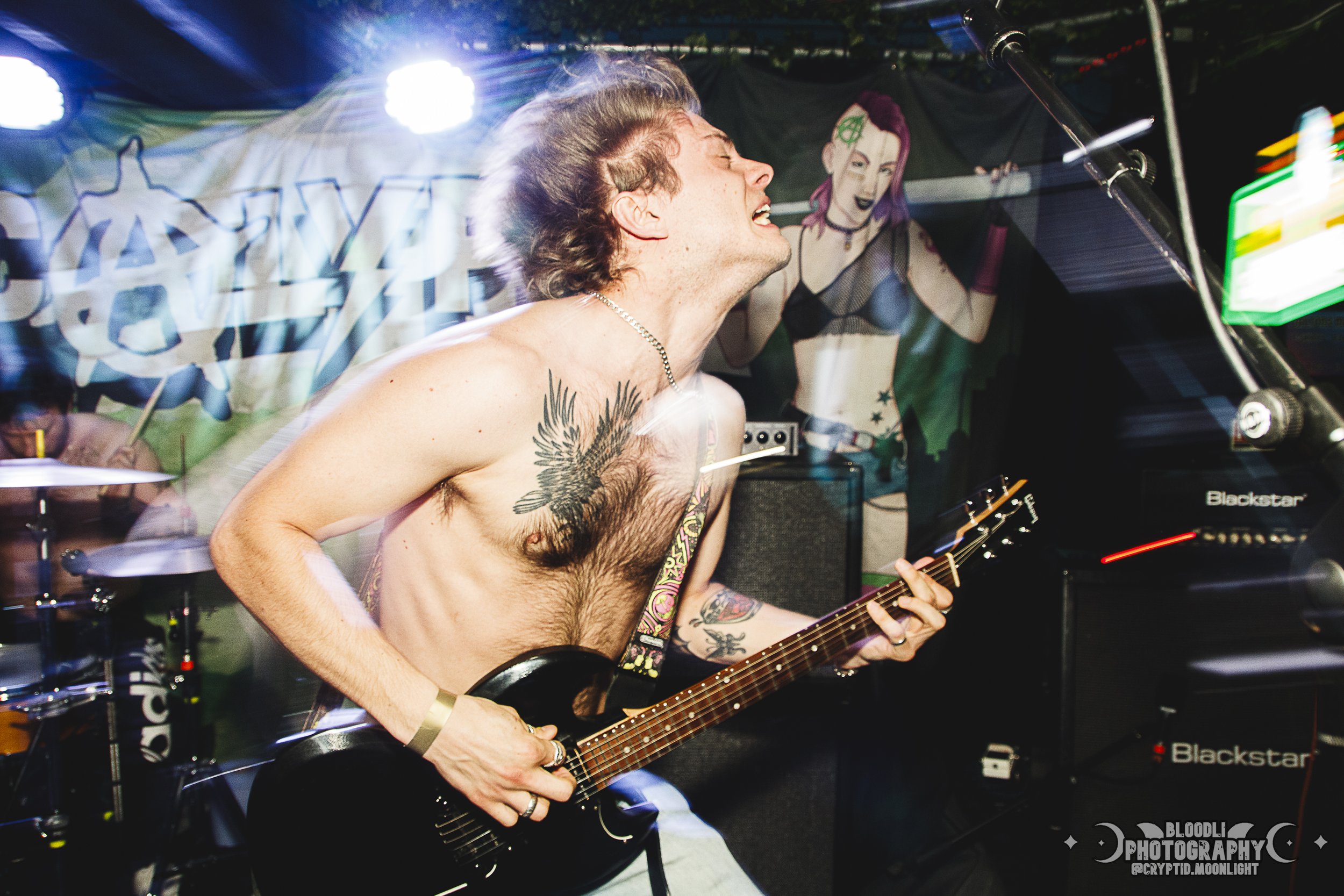 A shirtless male guitarist with tattoos on his chest and arm passionately playing an electric guitar during a live music performance at a concert or music event.