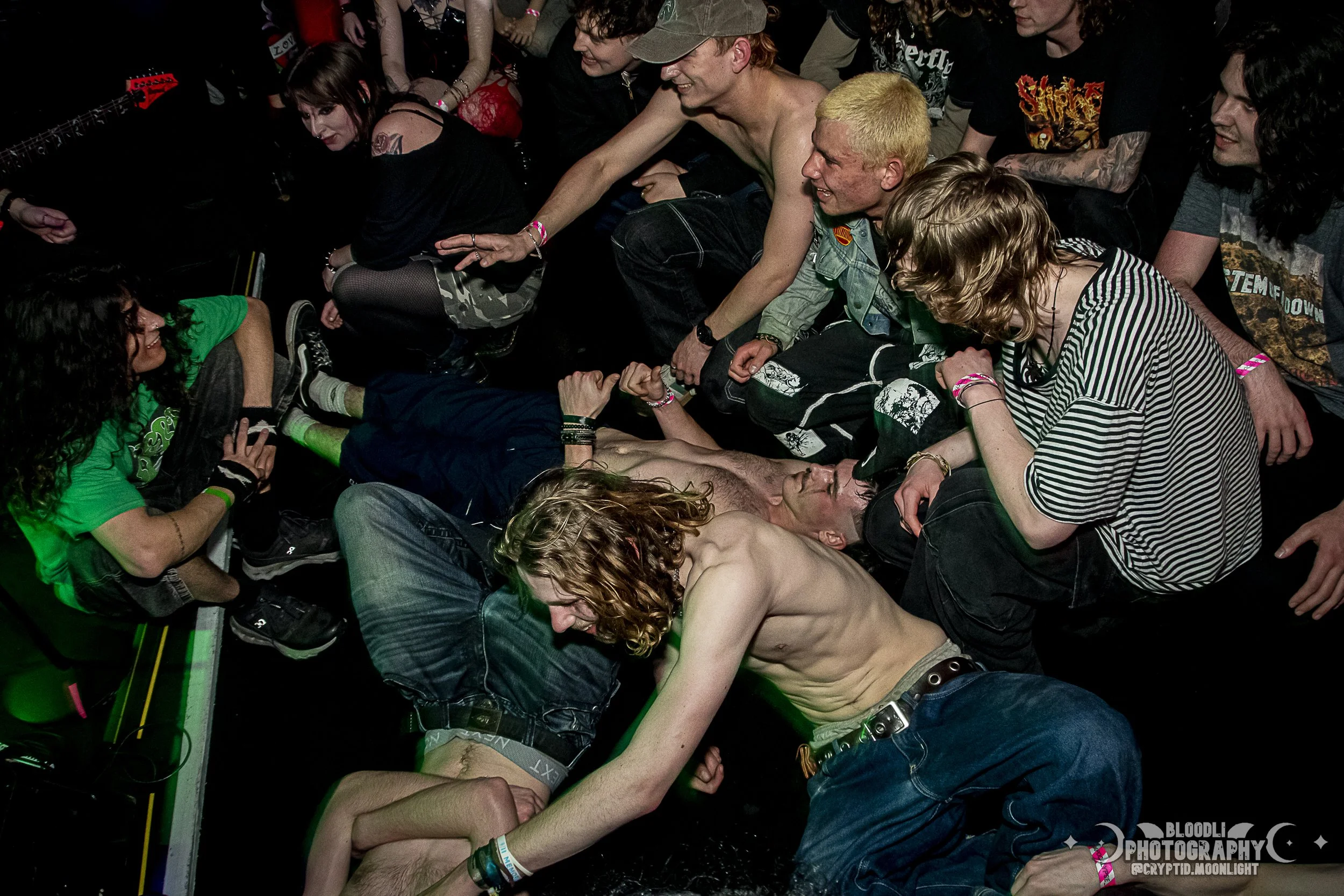 A crowded concert scene with a shirtless male performer lying on the floor, surrounded by a diverse group of people sitting and interacting with him.