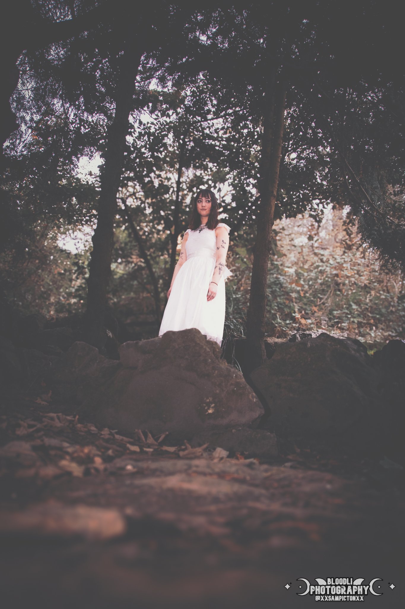 A woman in a white dress standing outdoors on a rocky area at dusk, surrounded by trees with an overcast sky.