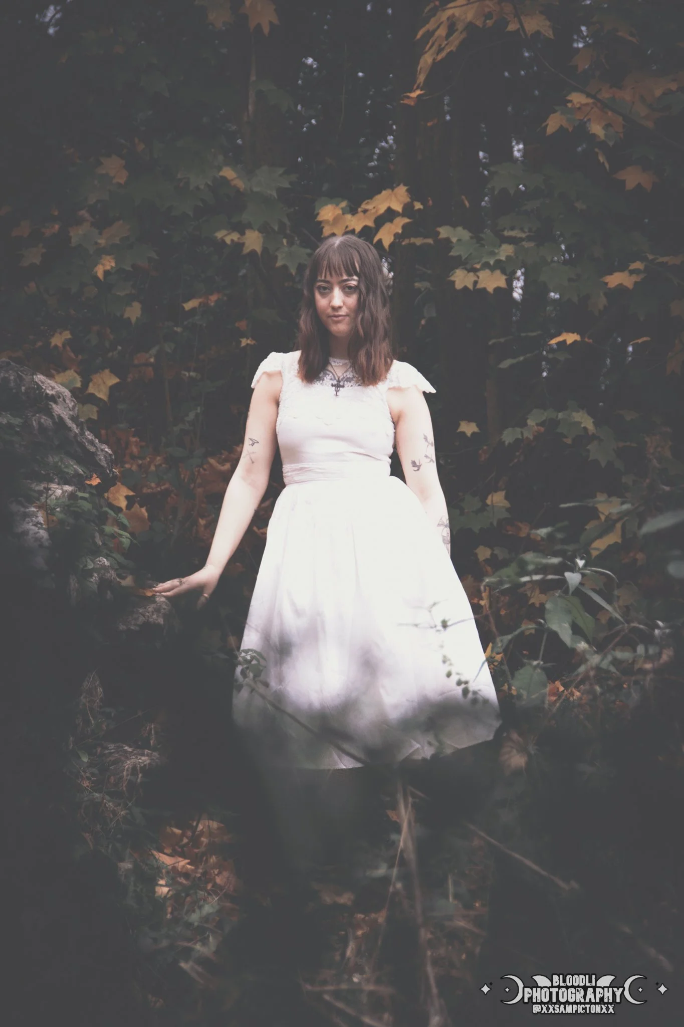 A young woman with wavy brown hair and tattoos on her arms stands outdoors in front of a background of green and brown autumn leaves, wearing a white dress. She is looking directly at the camera with a calm expression.