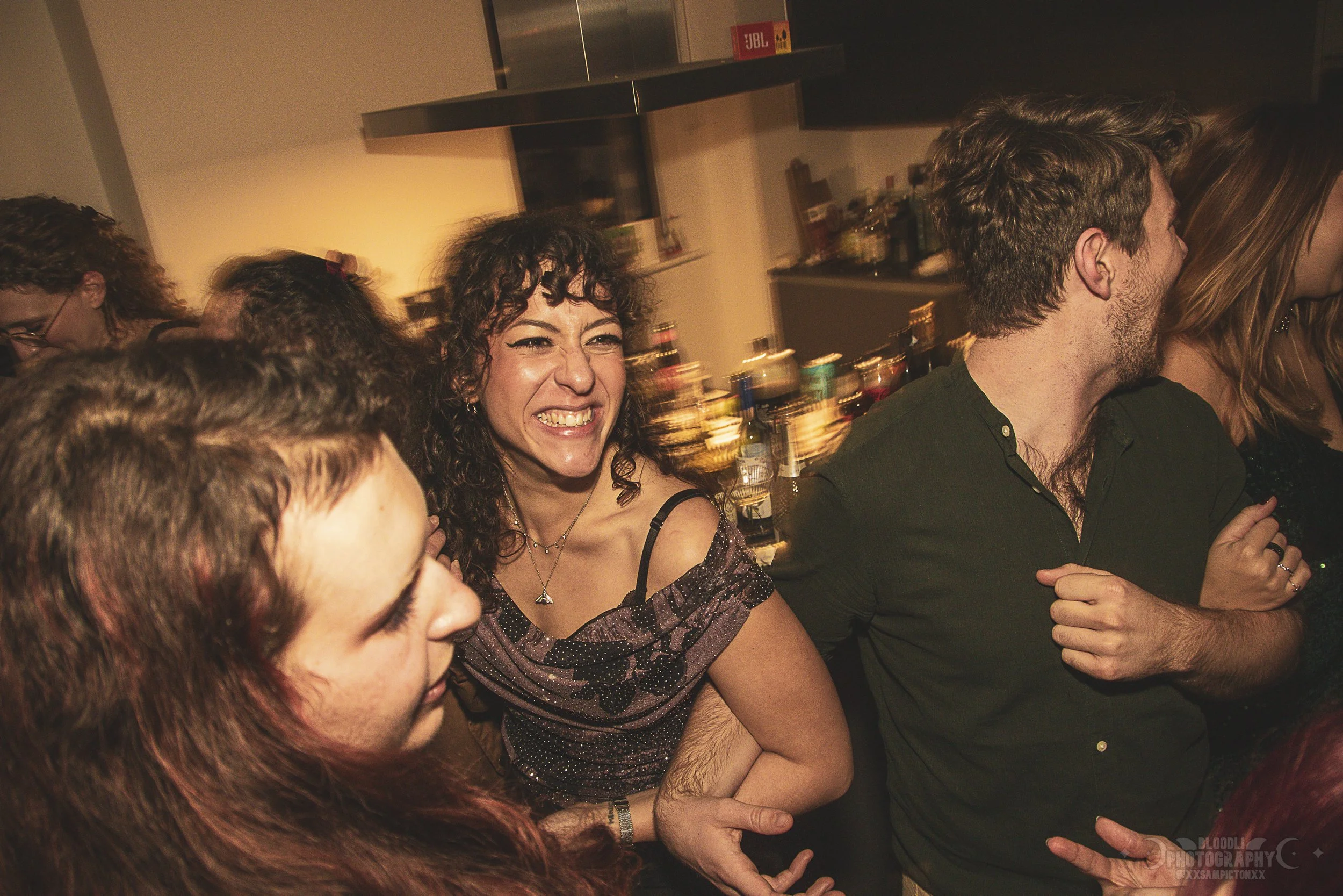 A group of people at a party, with a woman in the center smiling and laughing, surrounded by others, in a room with a bar in the background.