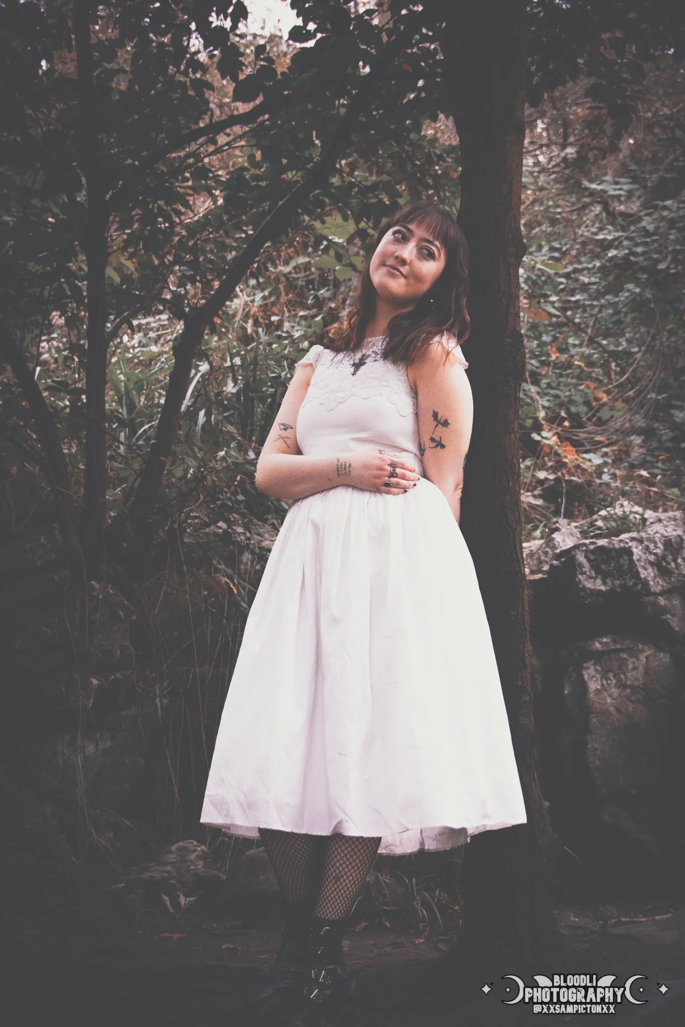 A young woman with tattoos on her arms and hands, wearing a white dress with lace details, fishnet stockings, and combat boots, standing in front of a tree in a forest or wooded area with dark foliage.