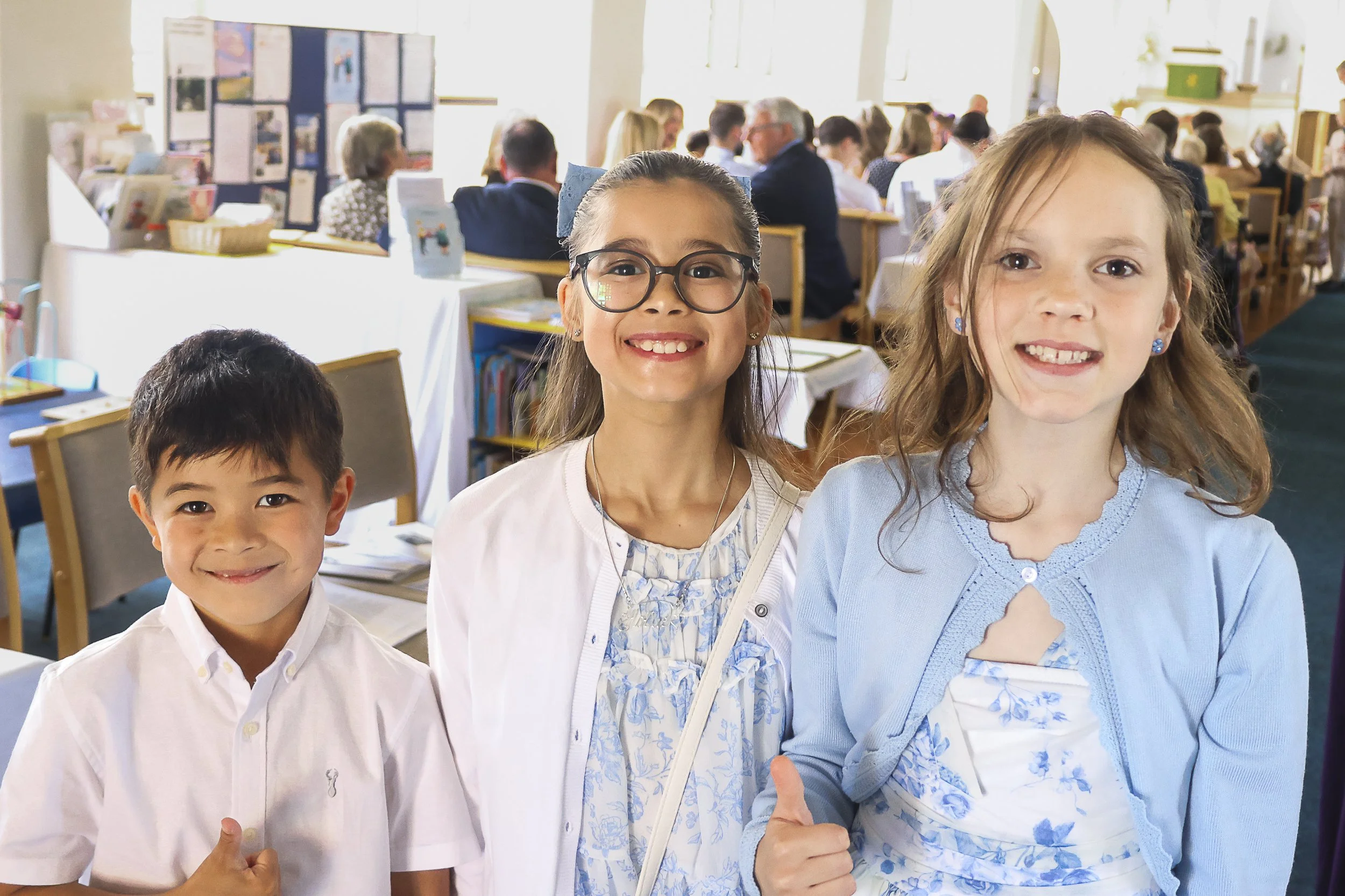 A group of three children smiling and giving thumbs up, standing in front of a lively indoor event with many people seated at tables.