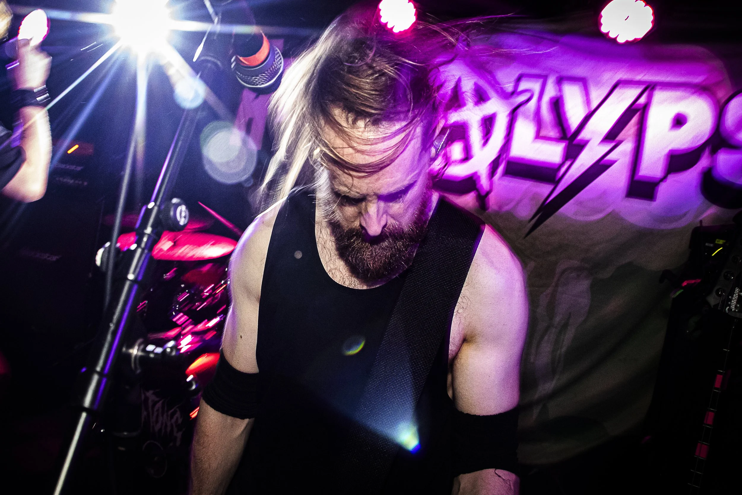A male musician with long hair and a beard performing live on stage, wearing a black sleeveless top, with purple and pink stage lighting and a banner with the word 'EXPLOSION' in the background.