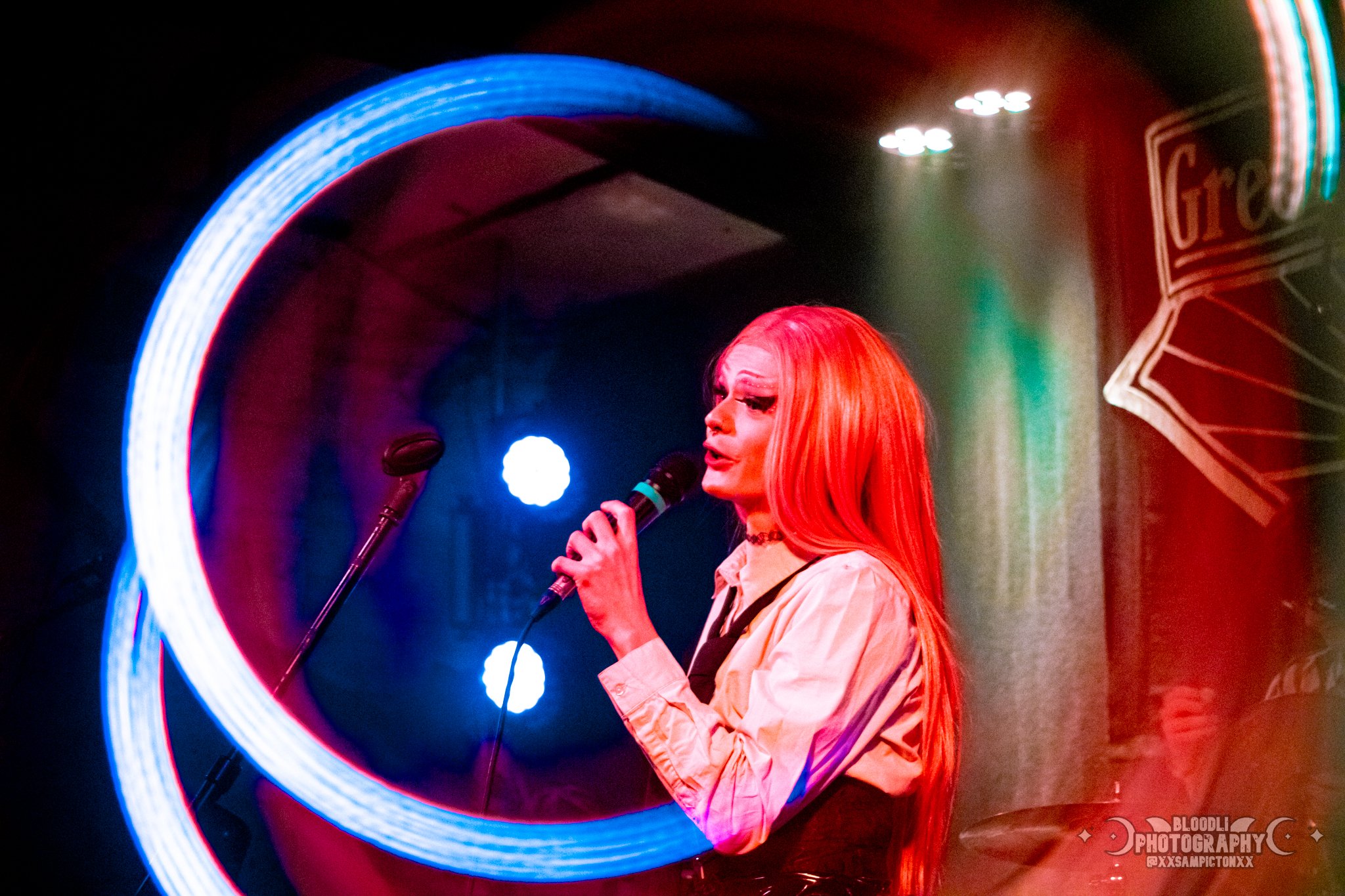 A woman with long red hair singing into a microphone on stage, illuminated by colorful stage lights with a neon sign in the background.