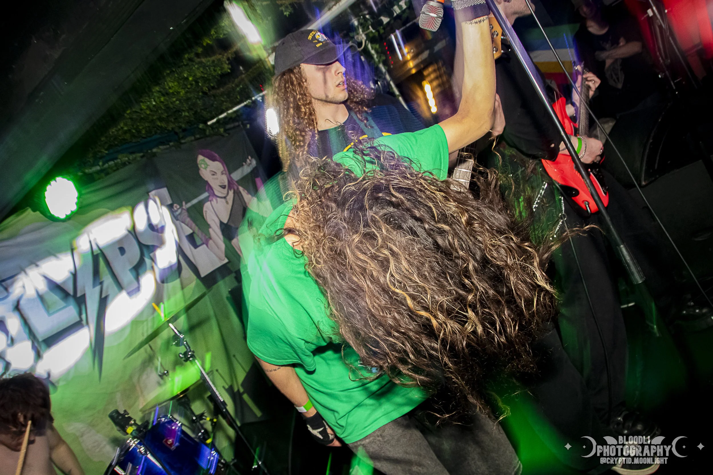 A musical band performing at a concert with green stage lighting. The band member with long curly hair wearing a green shirt is in the foreground, and another with long curly hair and a black cap is playing drums in the background. There is a colorfu
