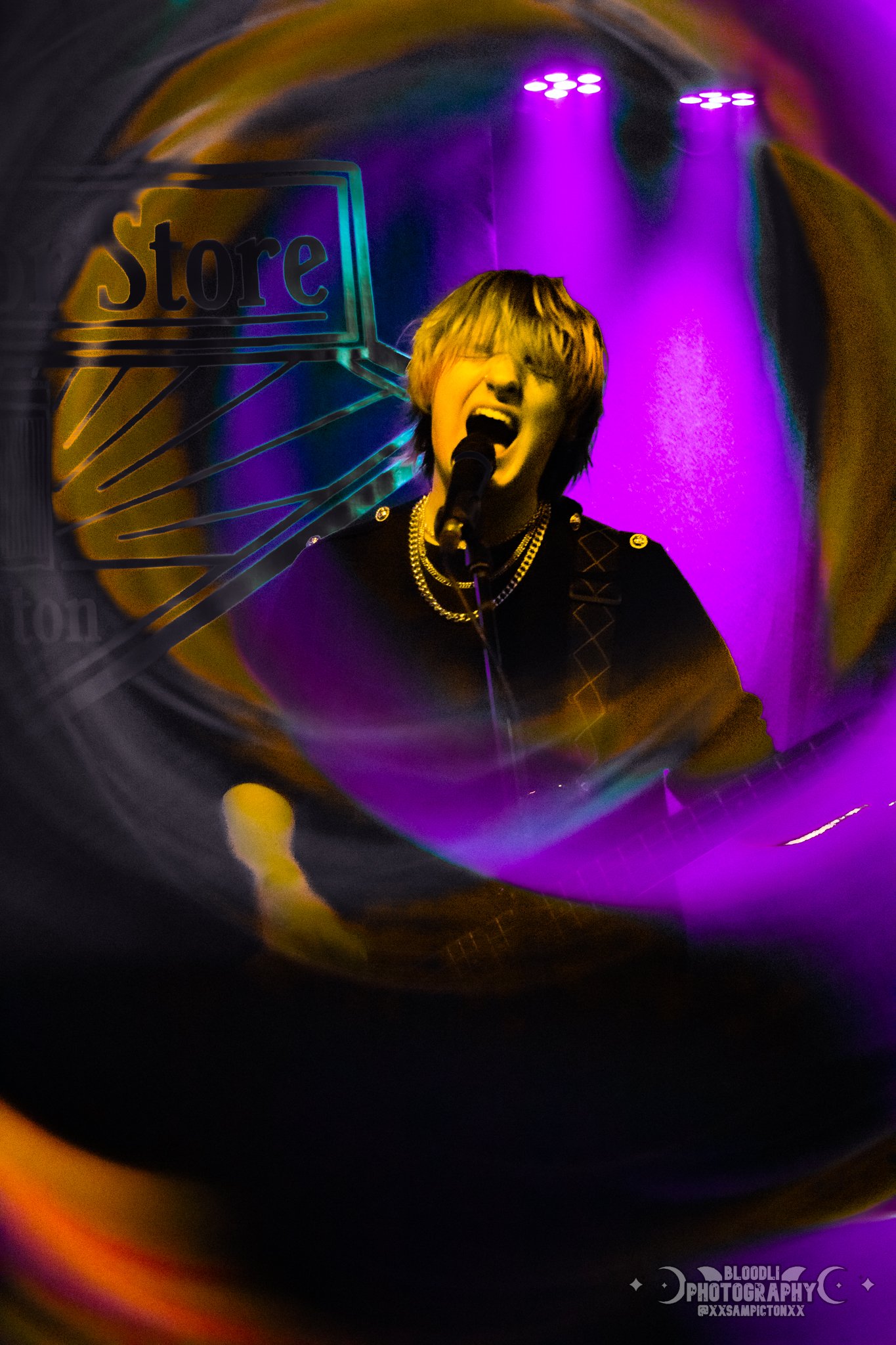 A young person singing into a microphone on stage, illuminated by purple and yellow stage lights, with a store sign visible in the background.