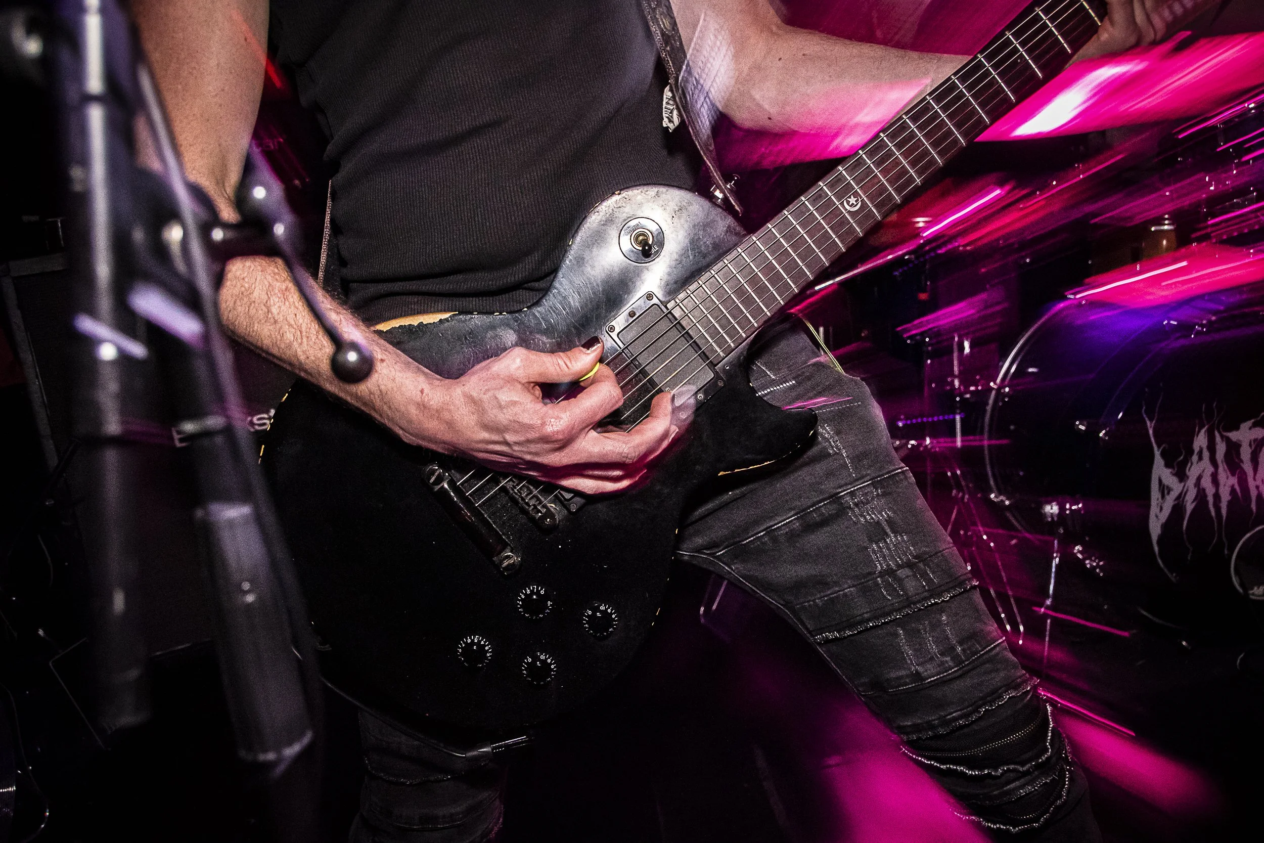 Person playing a black electric guitar with a dark shirt and ripped jeans in a dimly lit setting with pink and purple light streaks.