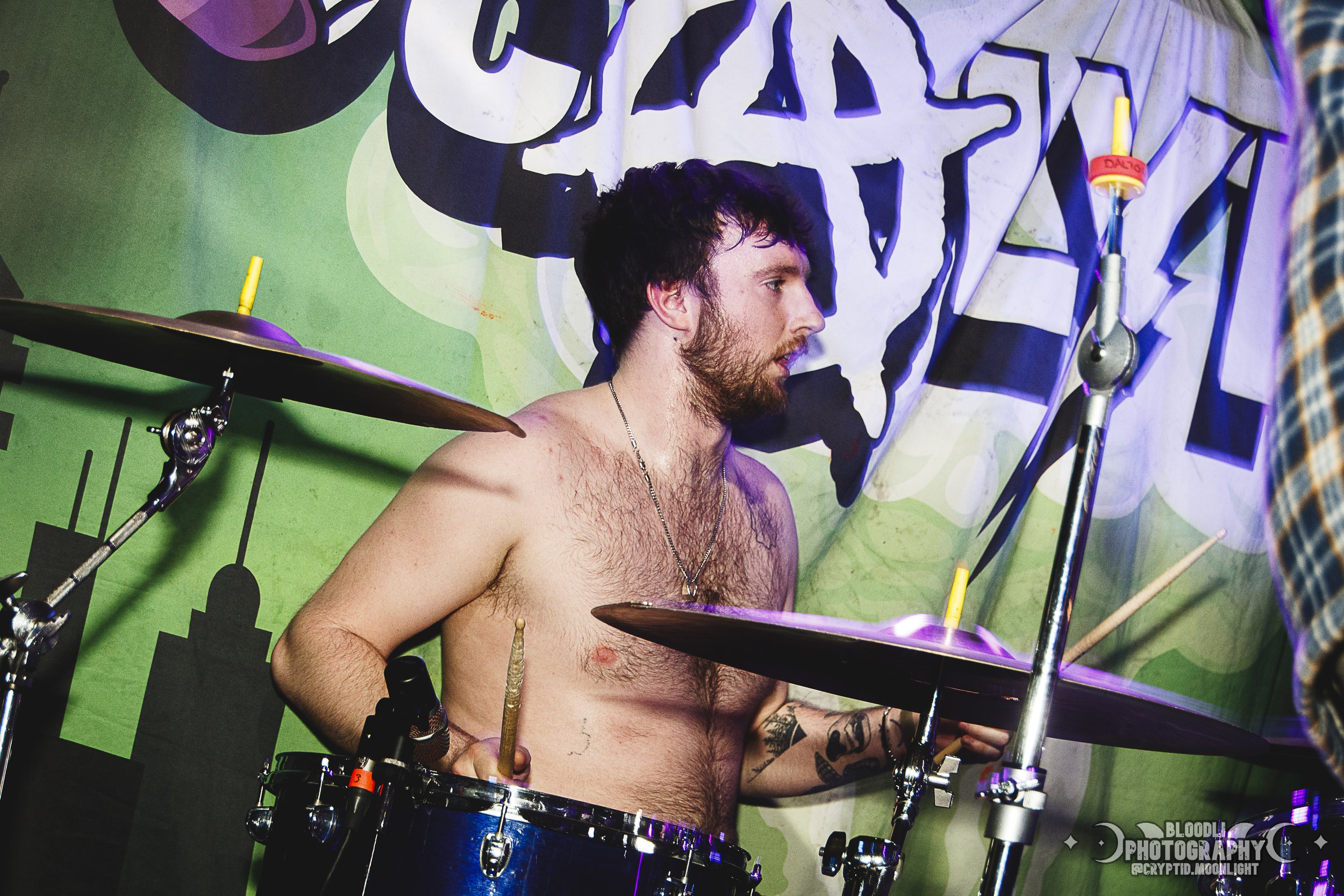 A shirtless man with a beard and tattoos playing the drums on a stage with a colorful backdrop.