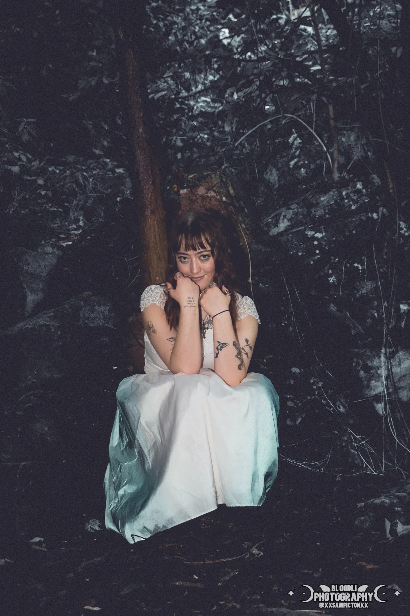 A woman with dark hair and tattoos, dressed in a white gown, sitting on the ground in a forest at night, with her arms resting on her knees and her chin resting on her hands, looking at the camera.
