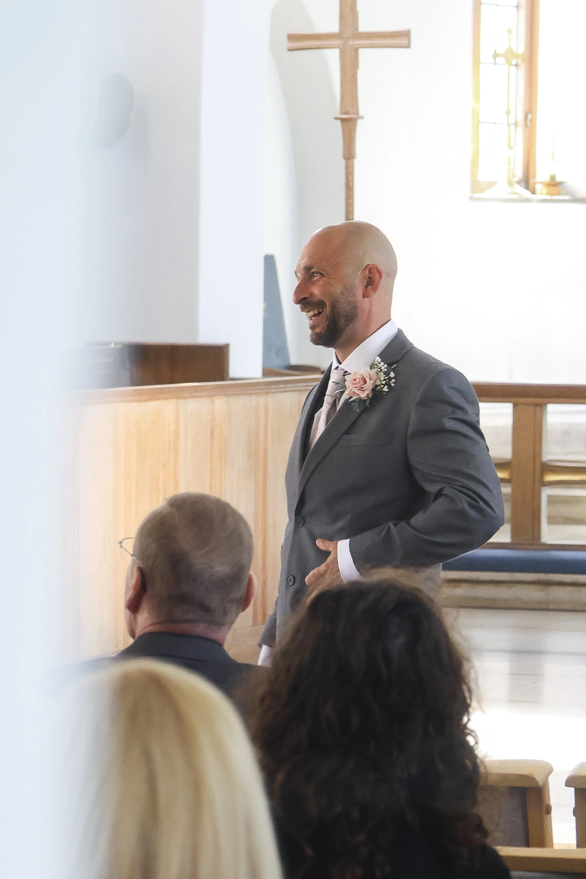 A man in a gray suit with a pink boutonniere is smiling during a ceremony at a church. There are people seated in the pews, and a wooden cross is in the background.