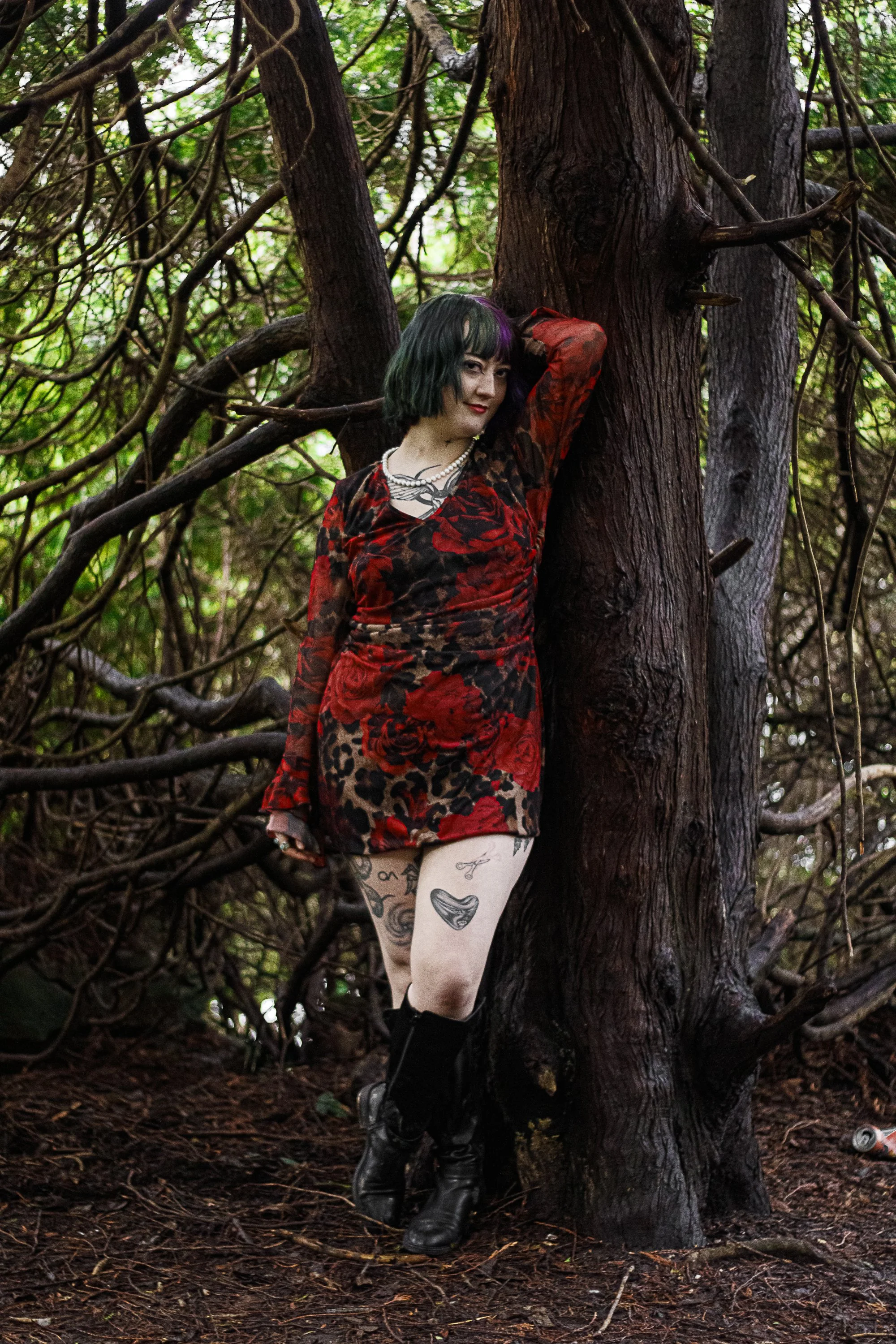 A woman with black and green hair, wearing a red and black floral and leopard print dress, standing in a forest, leaning against a large tree with tangled branches, with visible tattoos on her legs, wearing black boots, and jewelry, posing with one a