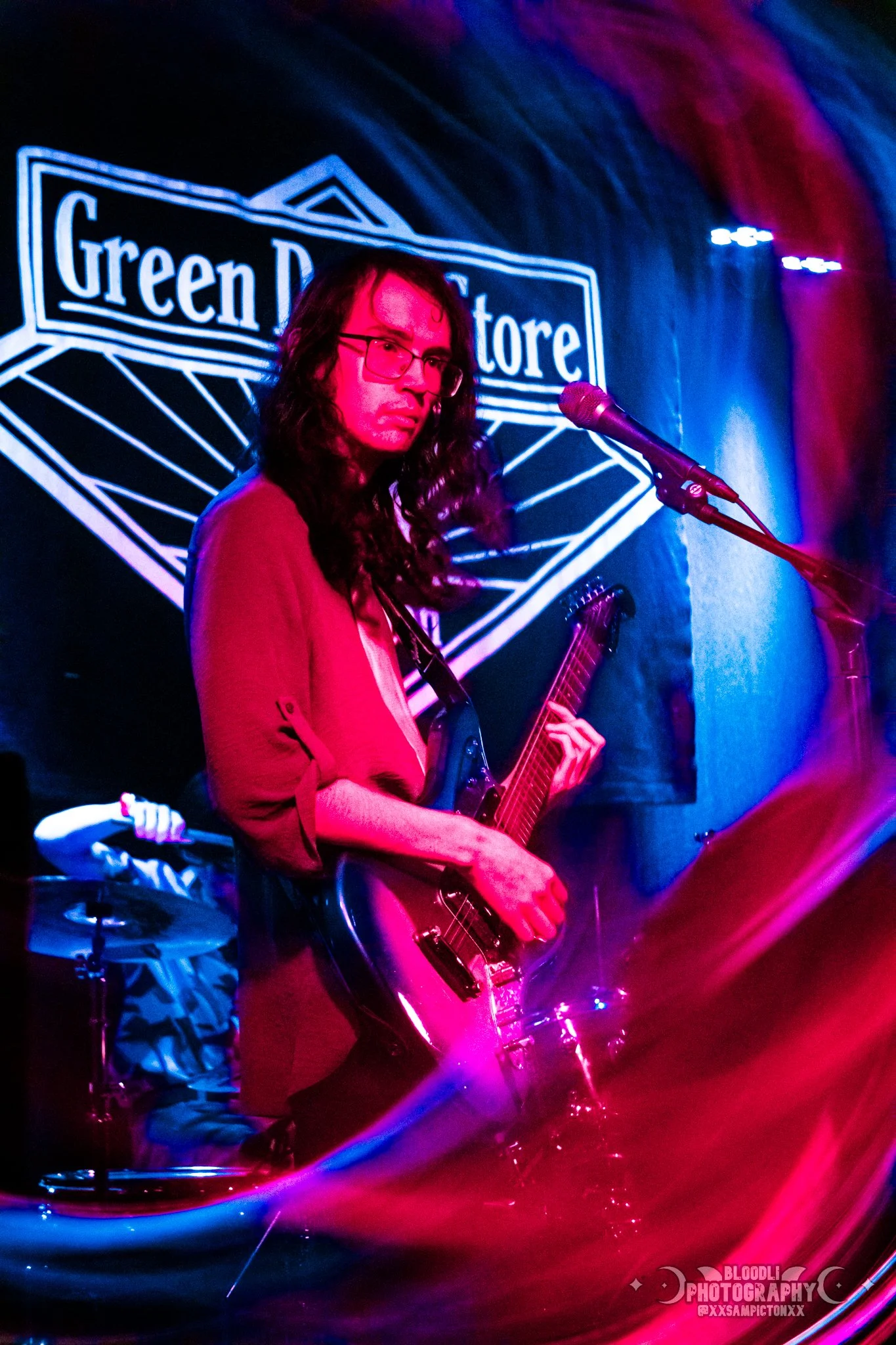 A woman playing an electric guitar on stage with colored stage lights, in front of a sign that reads 'Green Door Store'.