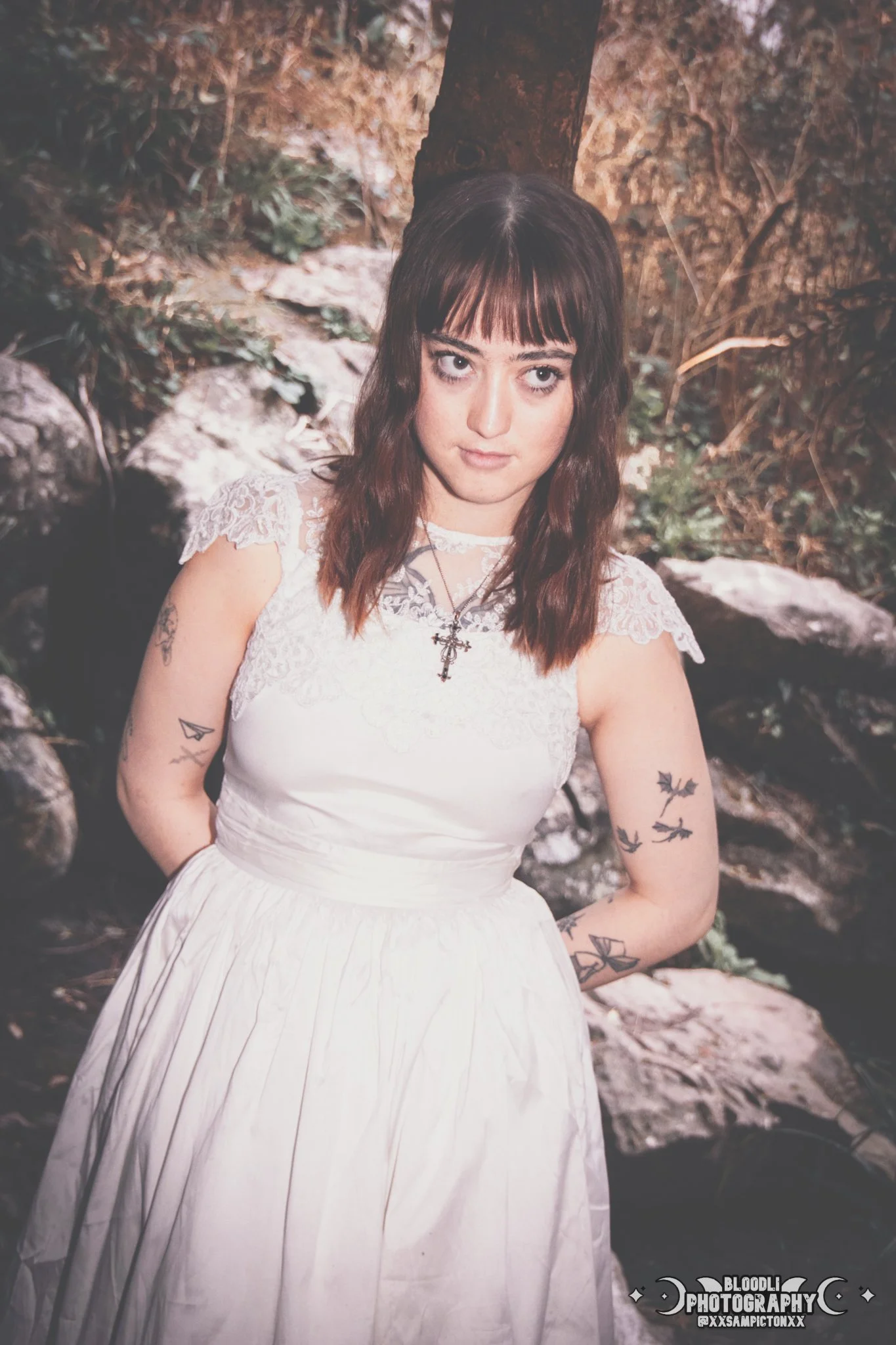 A young woman with brown hair and bangs stands outdoors in front of rocks and trees, wearing a white dress with lace on the sleeves, a cross necklace, and has tattoos on her arms.