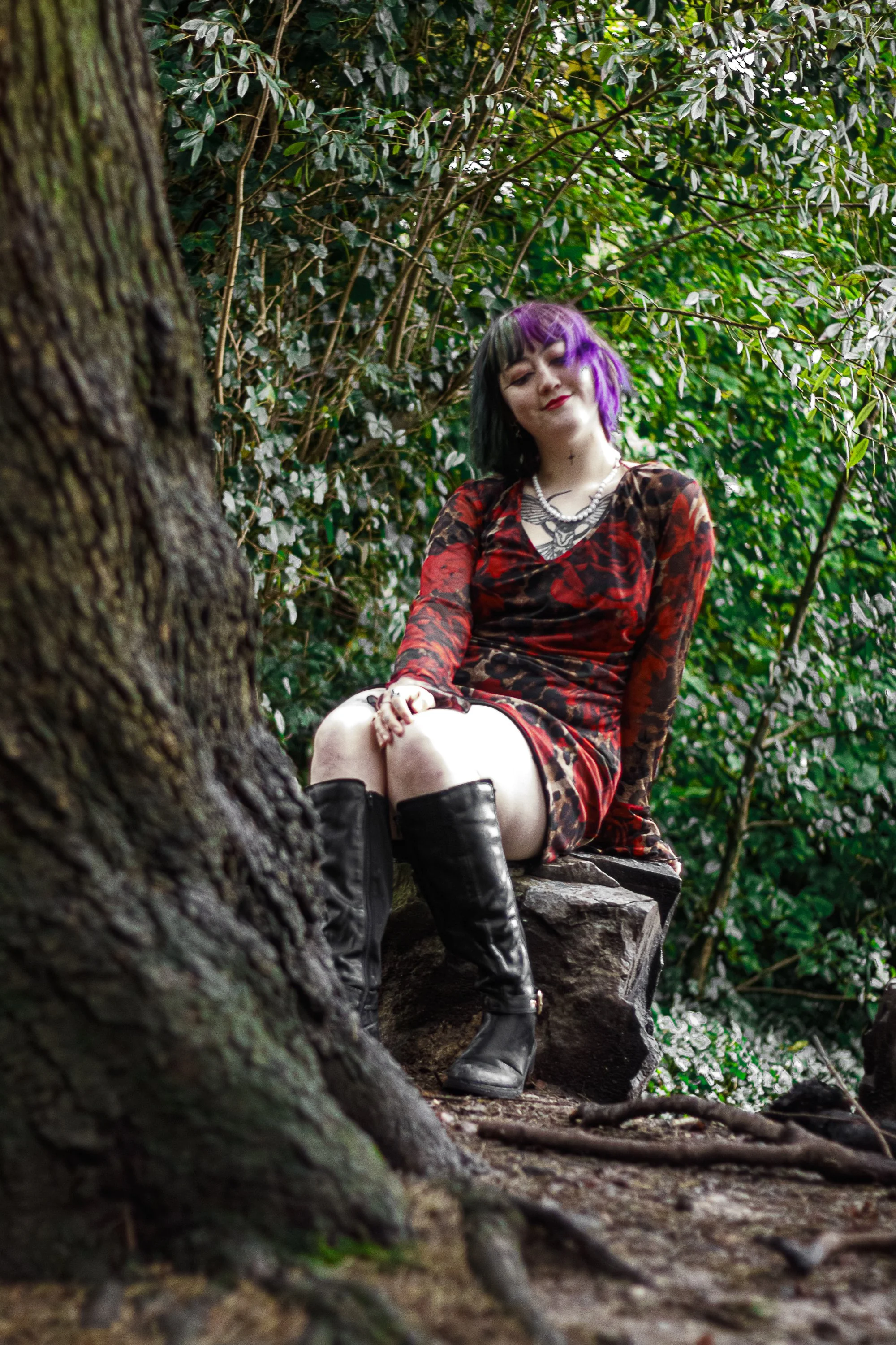 A woman with purple and black hair sitting on a rock in a wooded area, wearing a red and black dress and tall black boots.