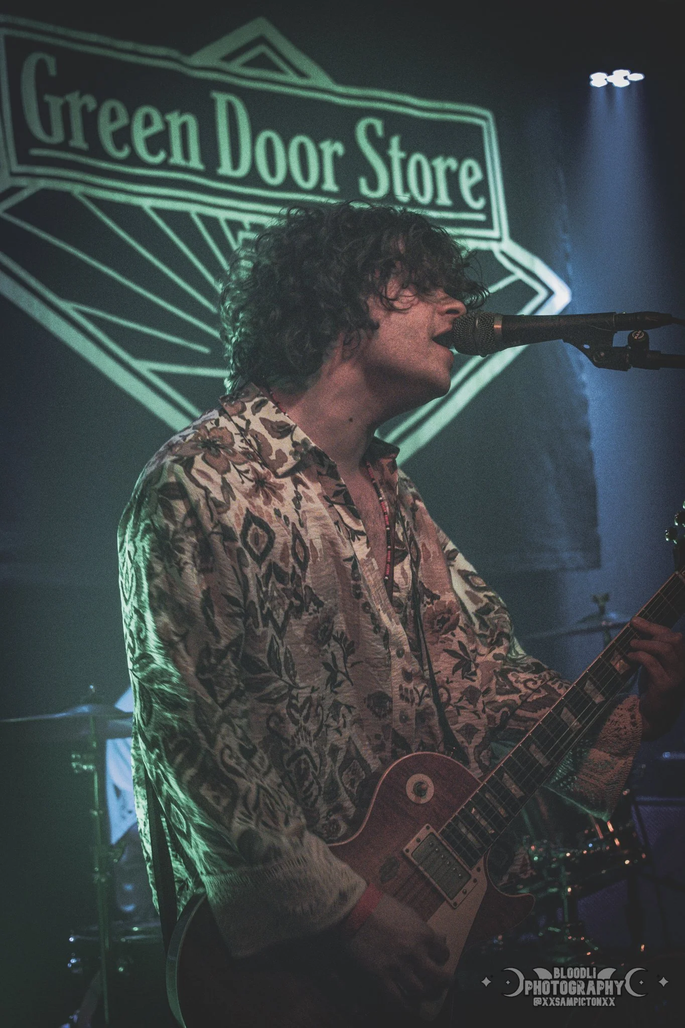 A person with curly hair singing into a microphone while playing an electric guitar on stage, with a sign that reads 'Green Door Store' in the background.