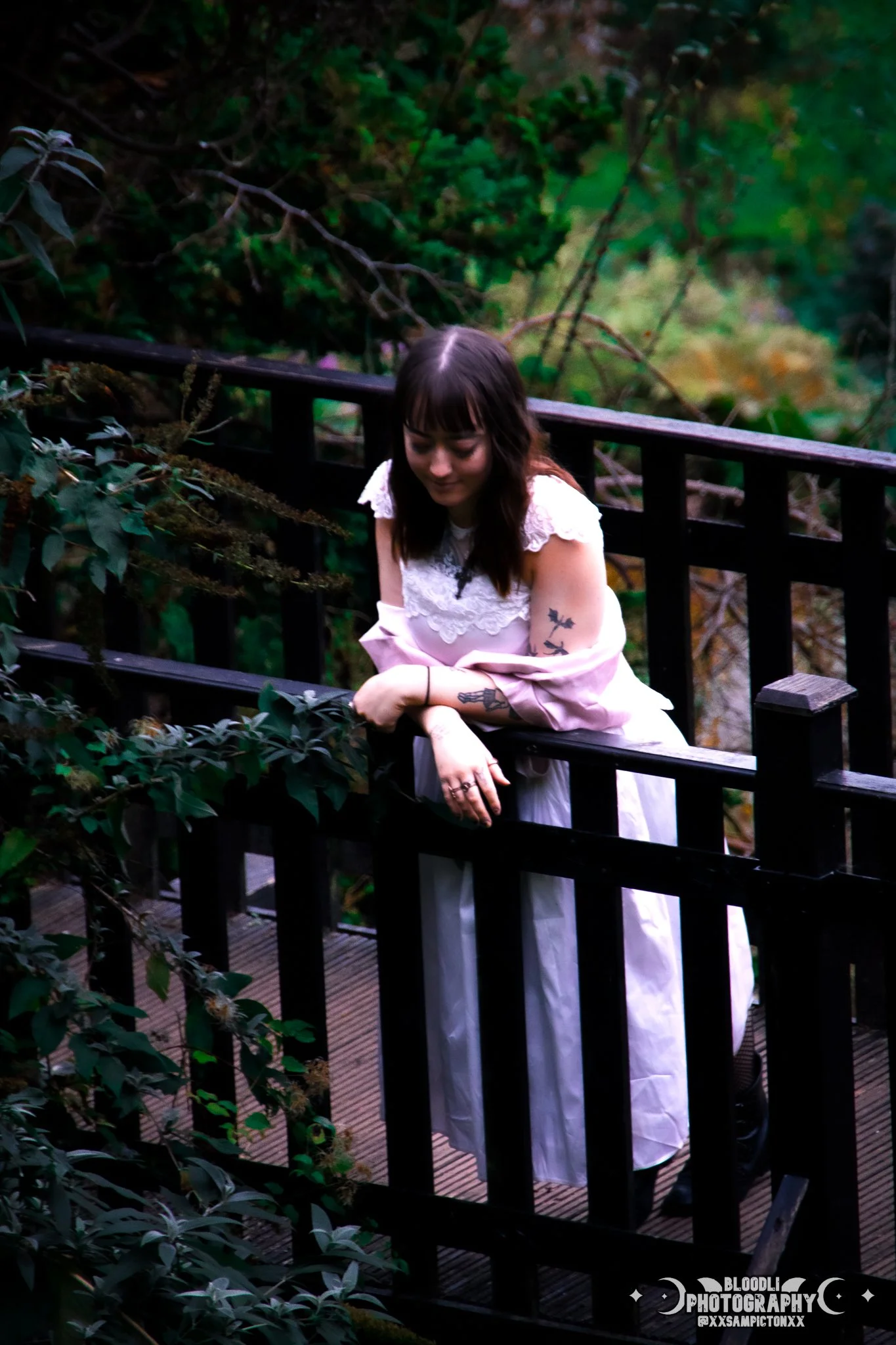 A young woman with tattoos, wearing a white dress and a light pink jacket, leaning on a black wooden railing on a bridge in a lush, green outdoor setting.