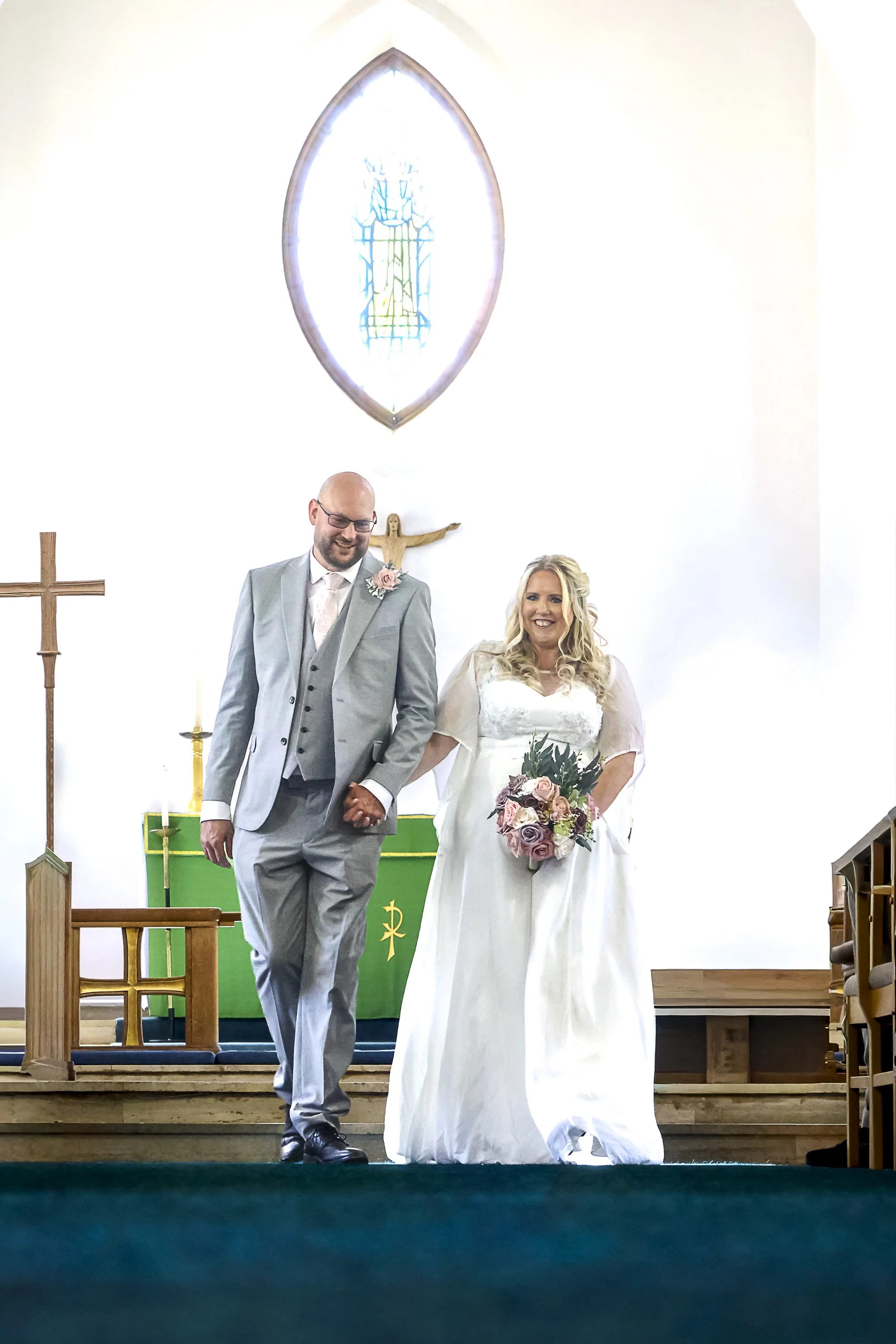 A bride and groom stand at the altar inside a church, holding hands, after a wedding ceremony. The groom is dressed in a light gray suit with a pink flower boutonniere, and the bride is wearing a white wedding dress and holding a bouquet of pink, pur