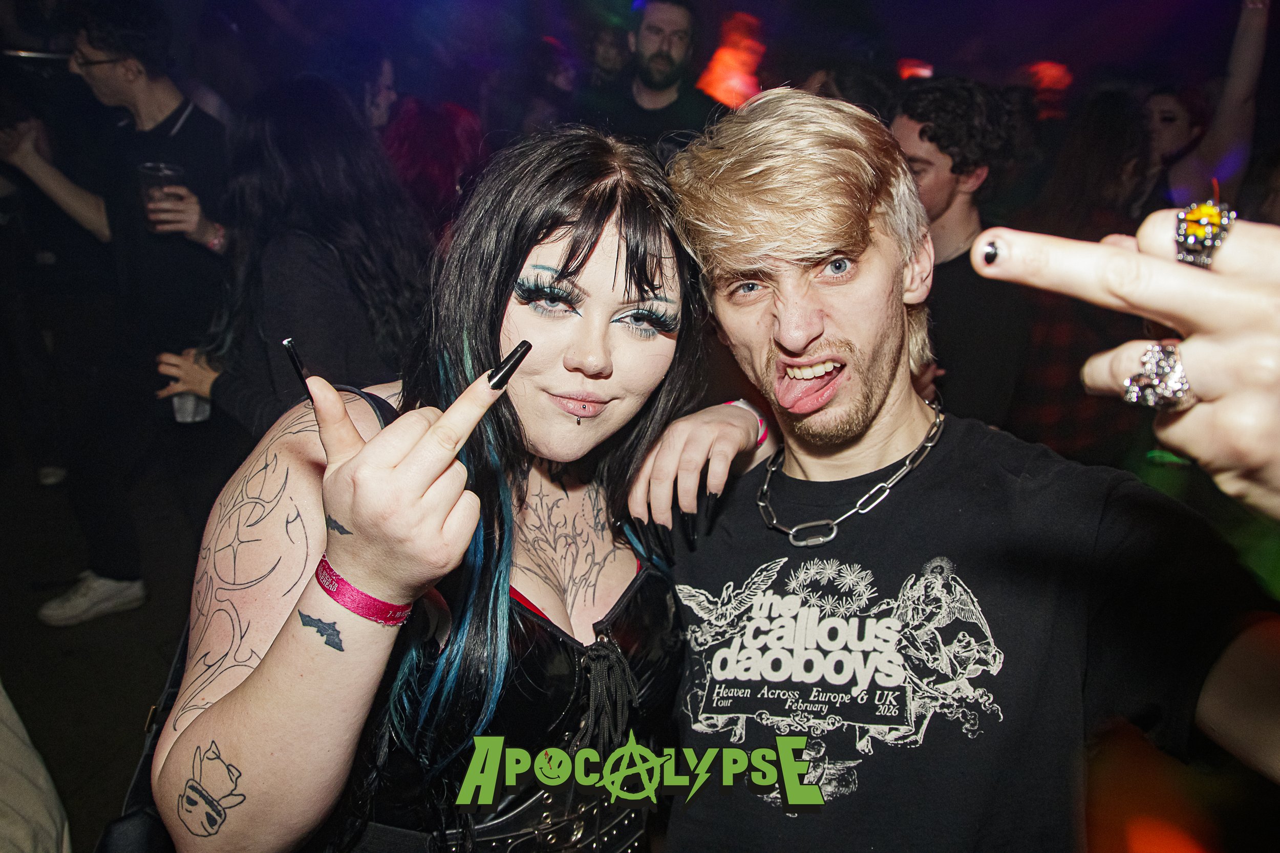 Two young adults at a concert or nightclub, making rude hand gestures. The woman has long black hair with blue streaks, tattoos on her arms, and wears a black top. The man has blonde hair, a nose ring, and wears a black t-shirt with band graphics. Th