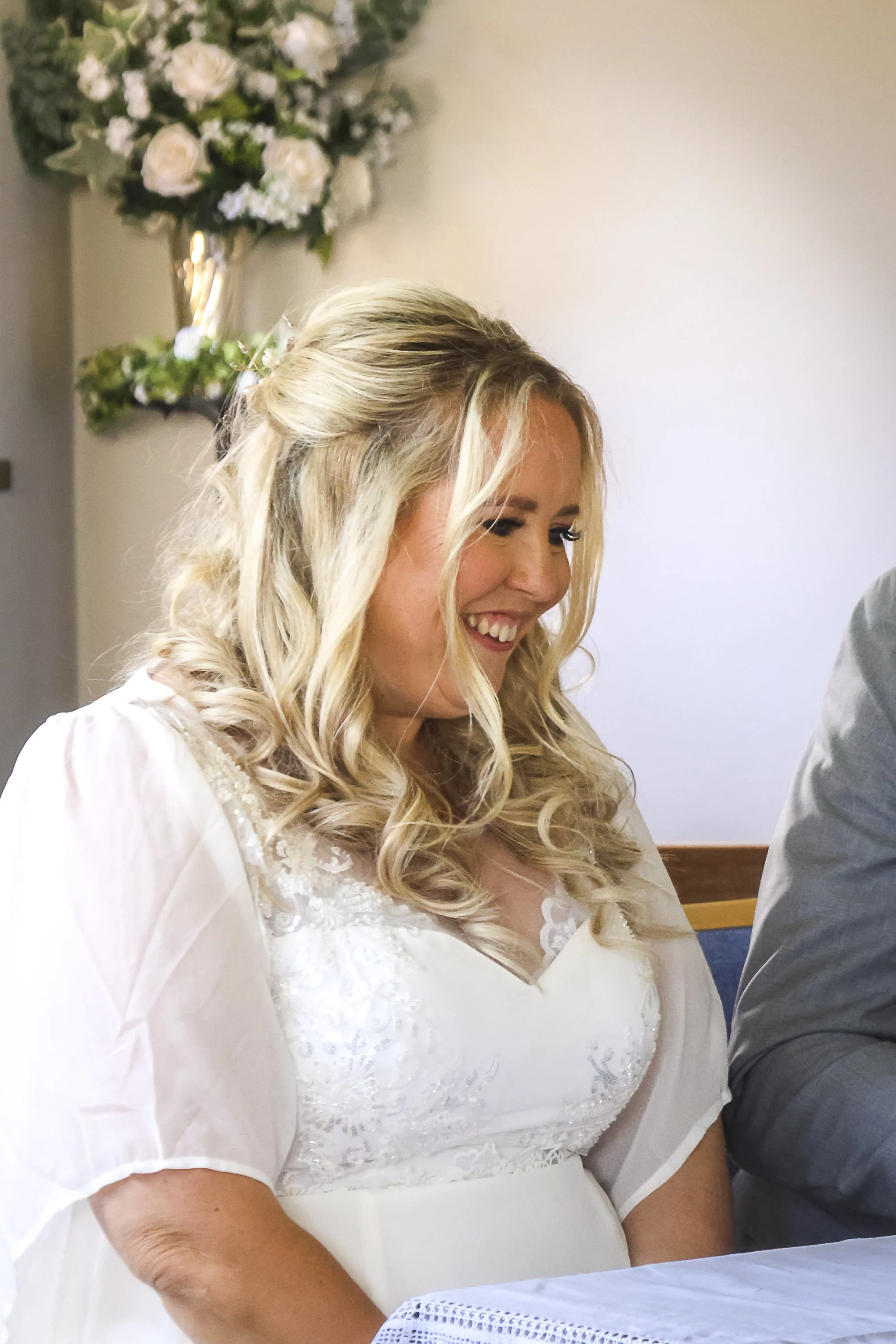 A woman with long blonde hair styled in loose curls, wearing a white lace wedding dress, smiling.