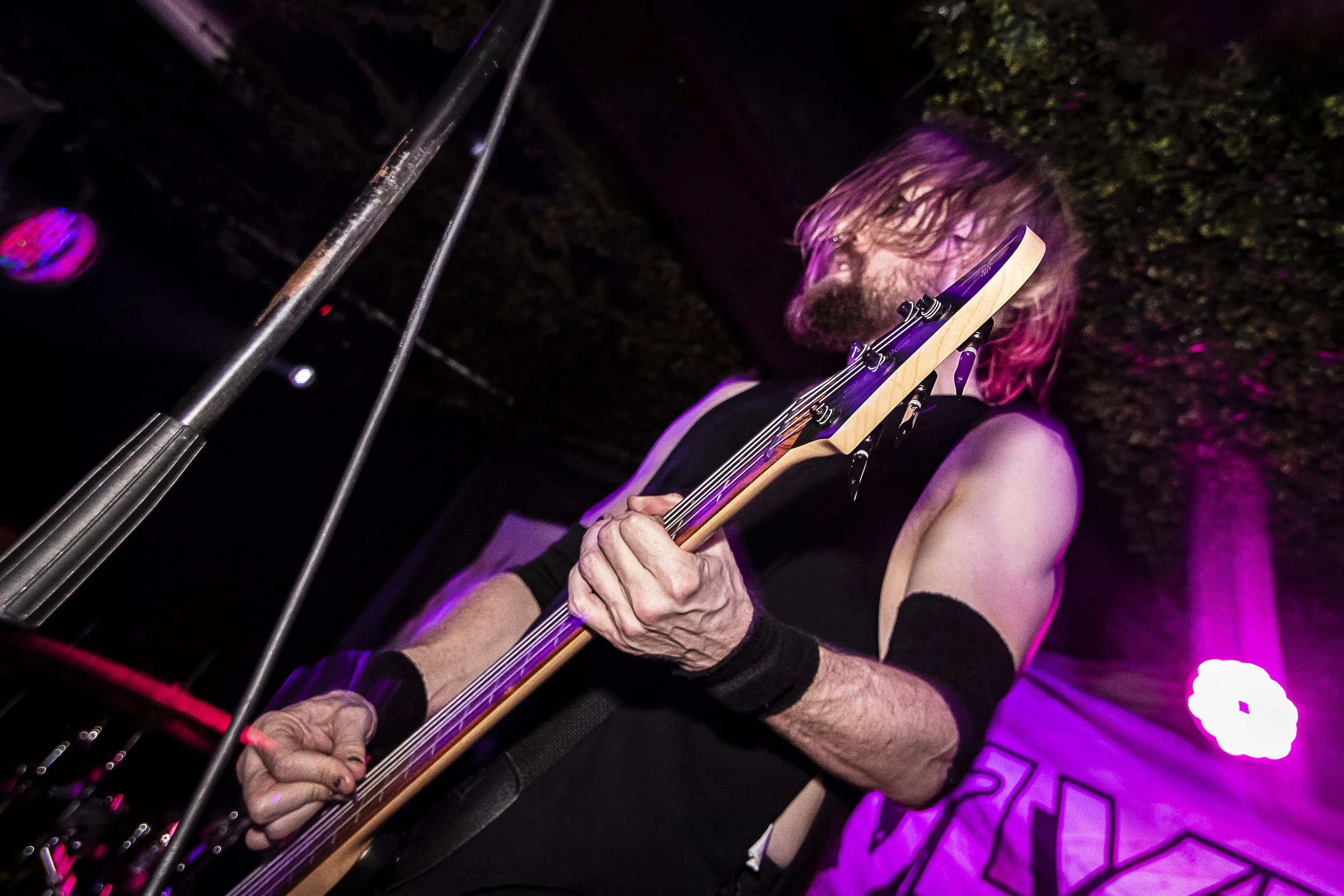 A musician with long hair and a beard playing an electric guitar on stage, illuminated by purple stage lights, with a poster or backdrop behind him.