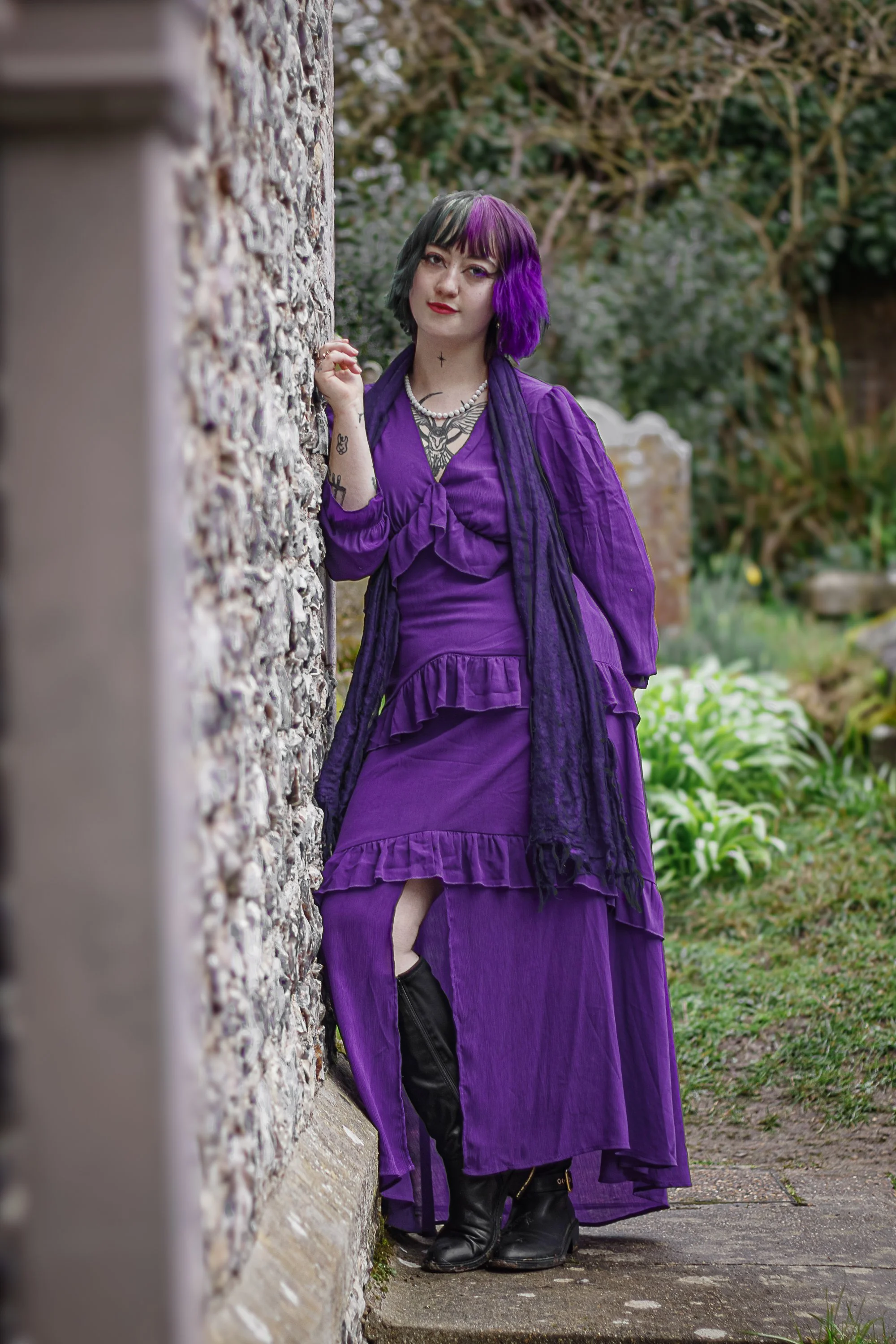A woman with black and purple hair stands outdoors, leaning against a textured stone wall. She is wearing a long purple dress with ruffles and a slit, paired with black thigh-high boots. She has tattoos and jewelry, and the background features greene