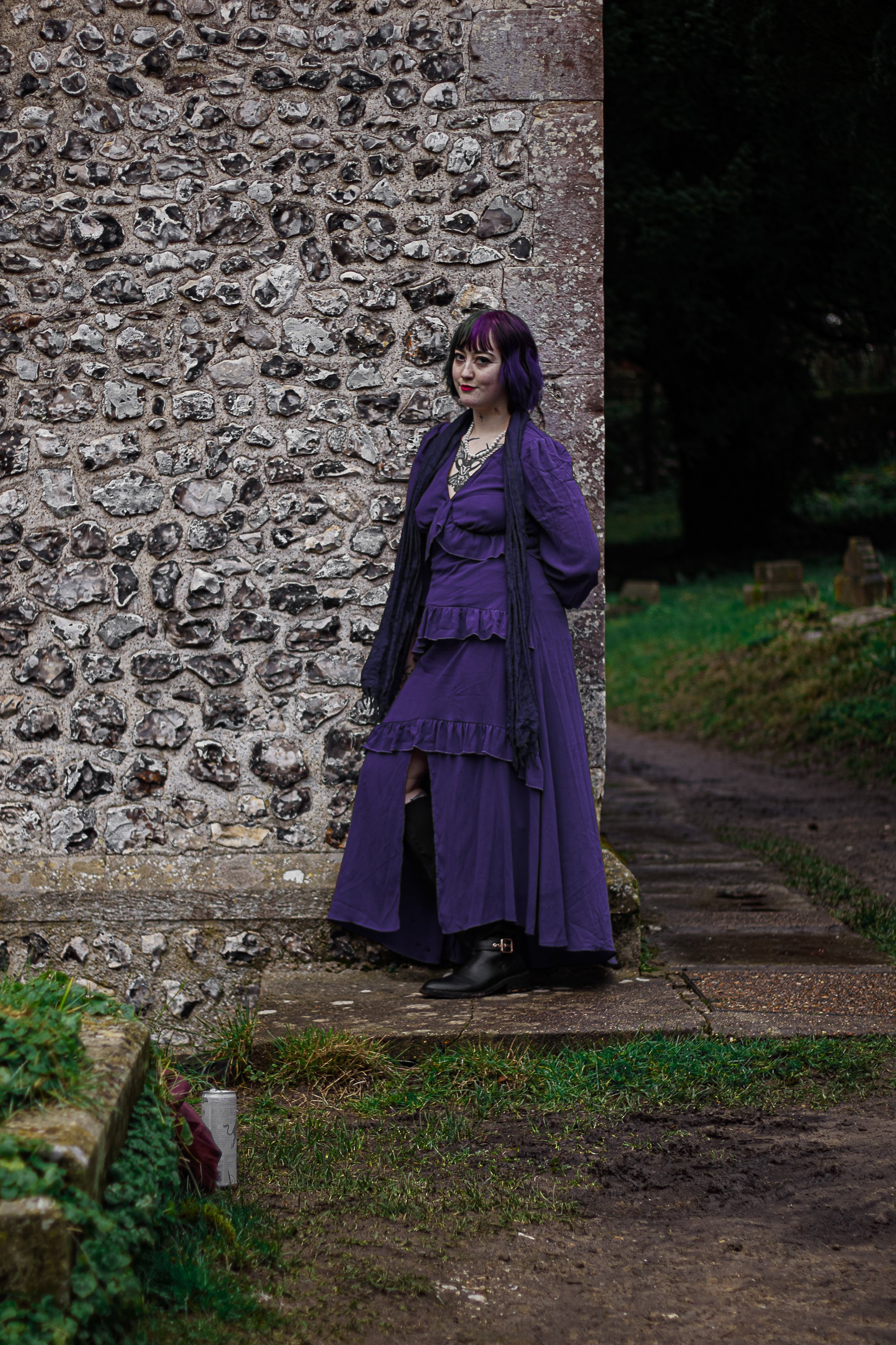A woman with purple and black hair, wearing a purple dress, black boots, and layered necklaces, standing outdoors against a stone wall
