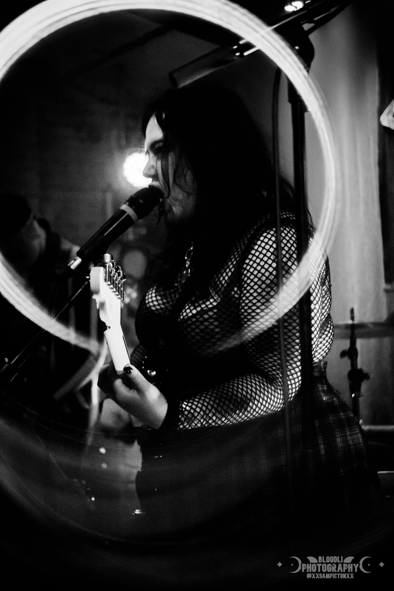 A woman singing into a microphone while playing an electric guitar, seen through a circular mirror or lens, in black and white.