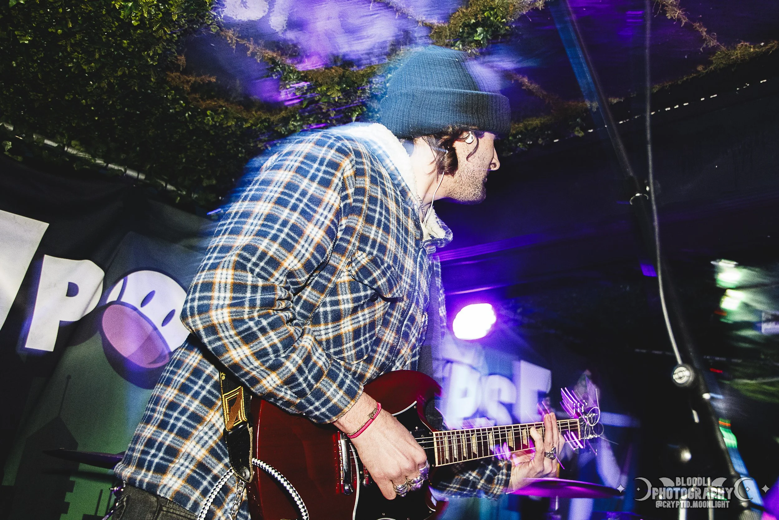 A musician wearing a black beanie and plaid shirt playing an electric guitar on stage at a concert.