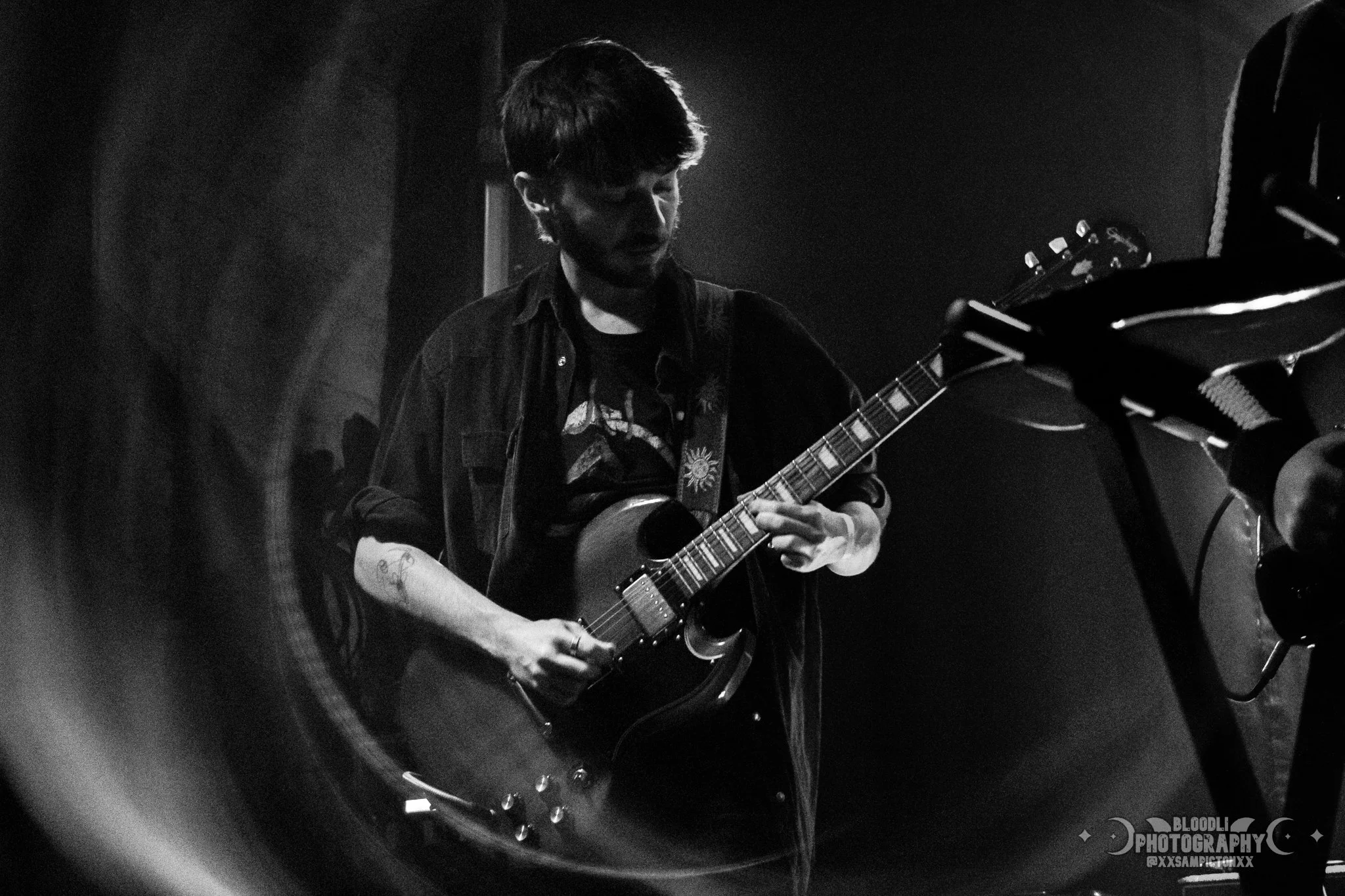 A young man playing an electric guitar on stage, in black and white, with a dark background and stage lighting.
