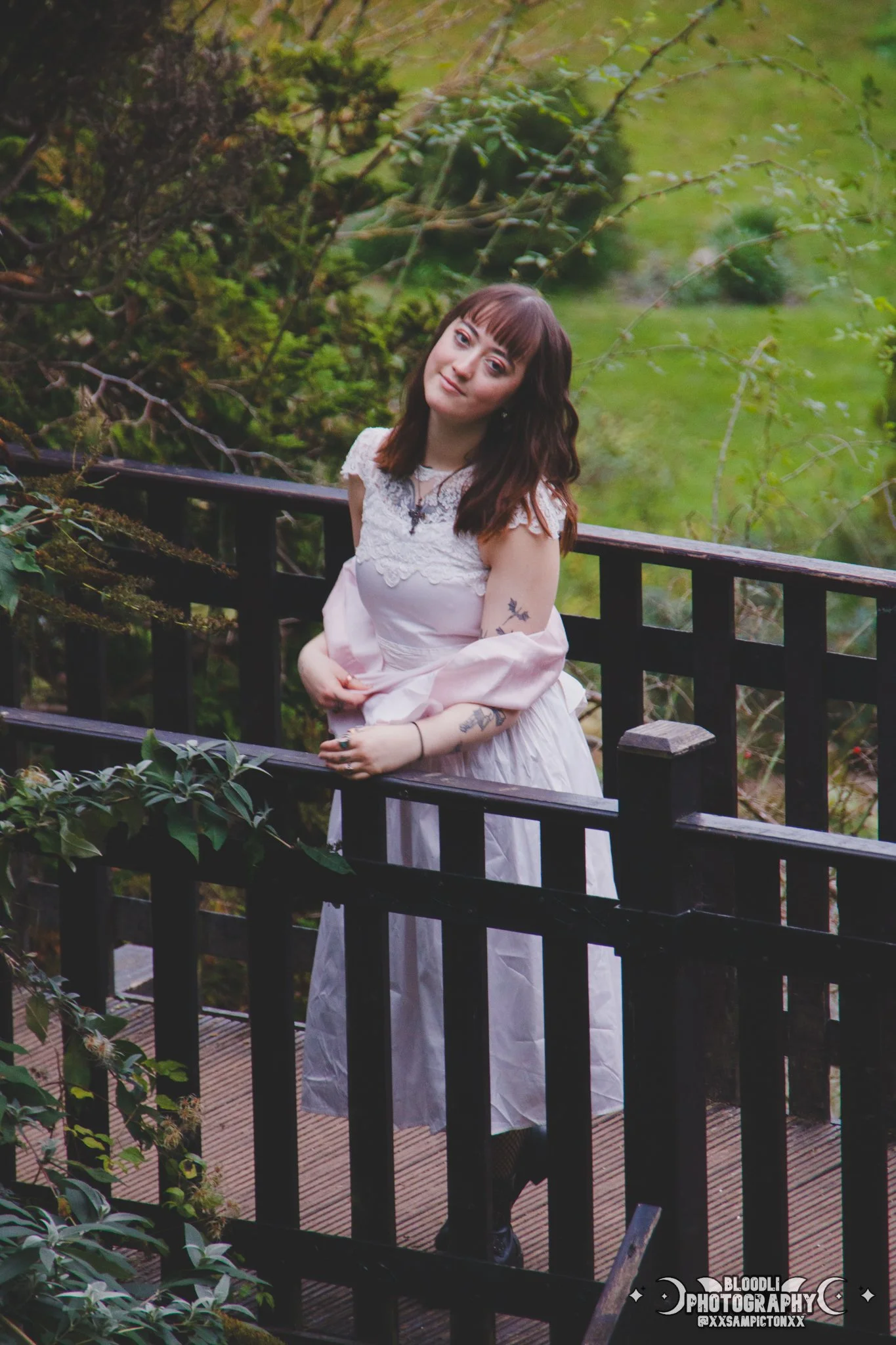 A woman with brown hair, wearing a white vintage-style dress with lace details, standing on a wooden bridge in a lush green forest. She has tattoos on her arms and is looking at the camera with a subtle smile.