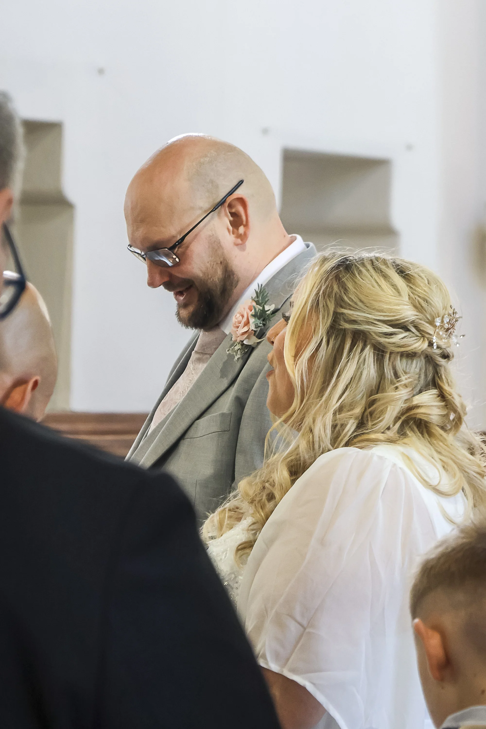 A man with a beard and glasses, dressed in a gray suit with a pink flower on his lapel, is smiling and looking downward during a wedding ceremony with a blonde woman in a white dress with lace details and a pearl hairpiece.