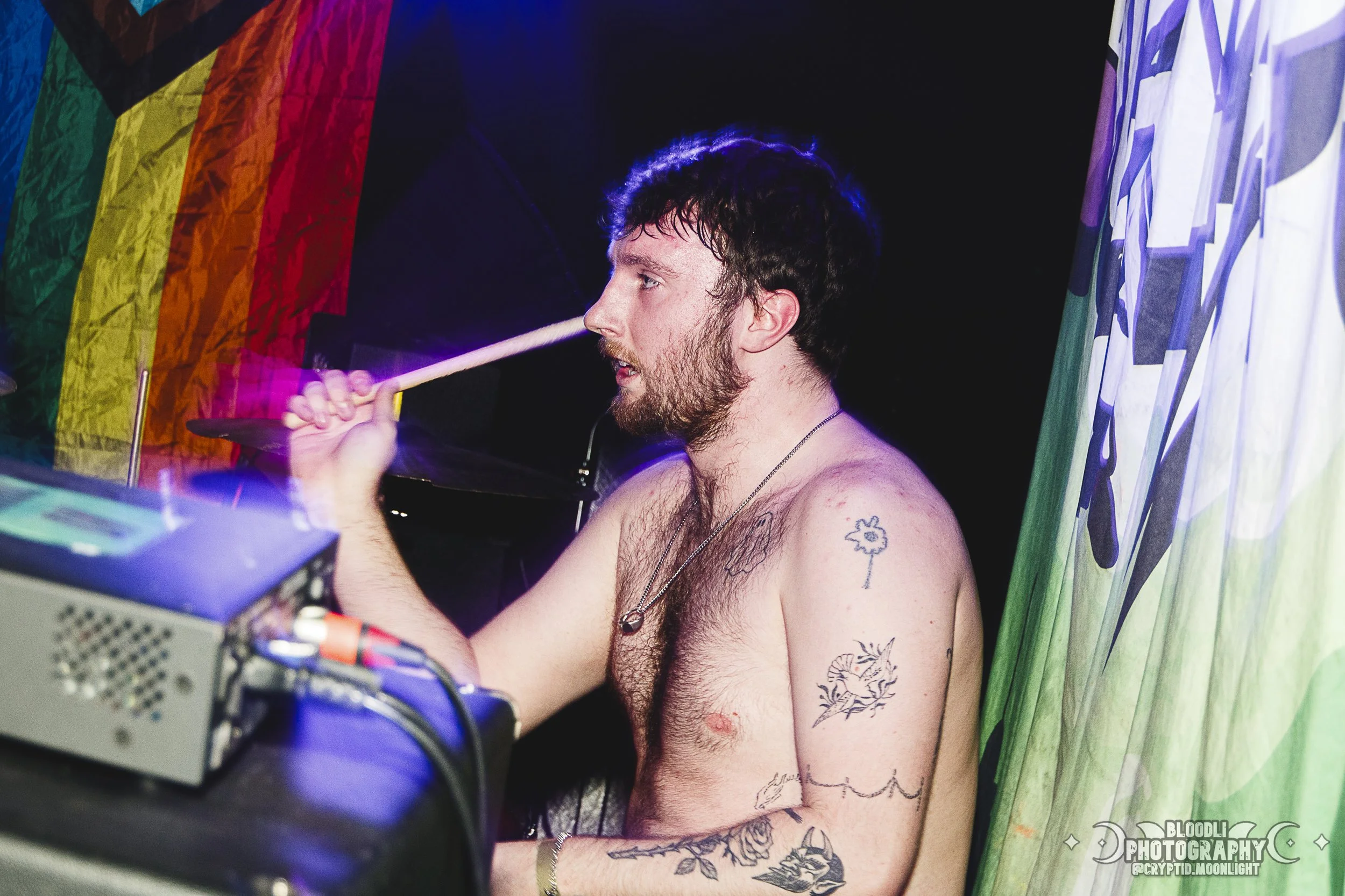 A shirtless man with tattoos on his arm and shoulder, holding a drumstick near his face, appears to be DJing or performing at an event with rainbow flags and colorful decorations in the background.