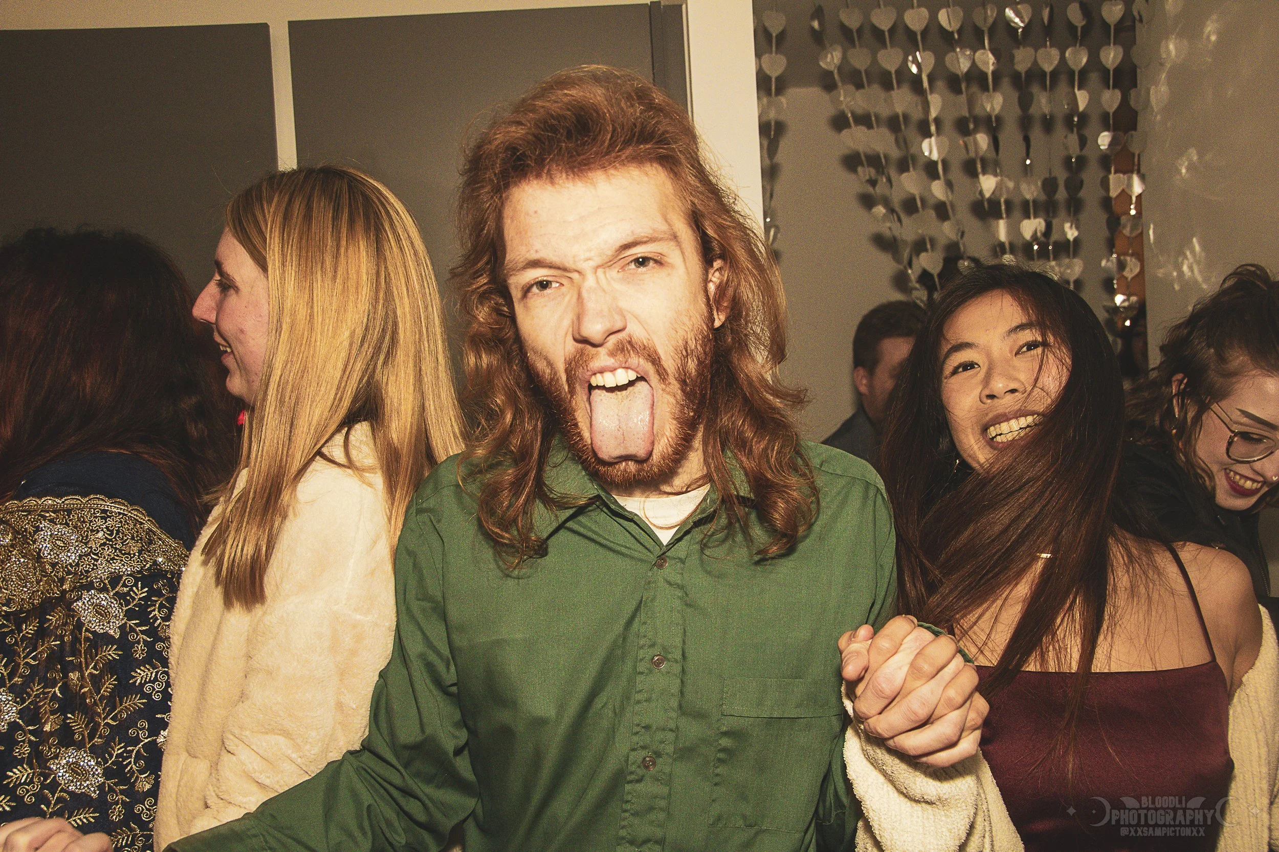 Group of people at party, central man with long hair, beard, and green shirt sticking out tongue, holding hands, smiling, other women in background wearing colorful clothing.
