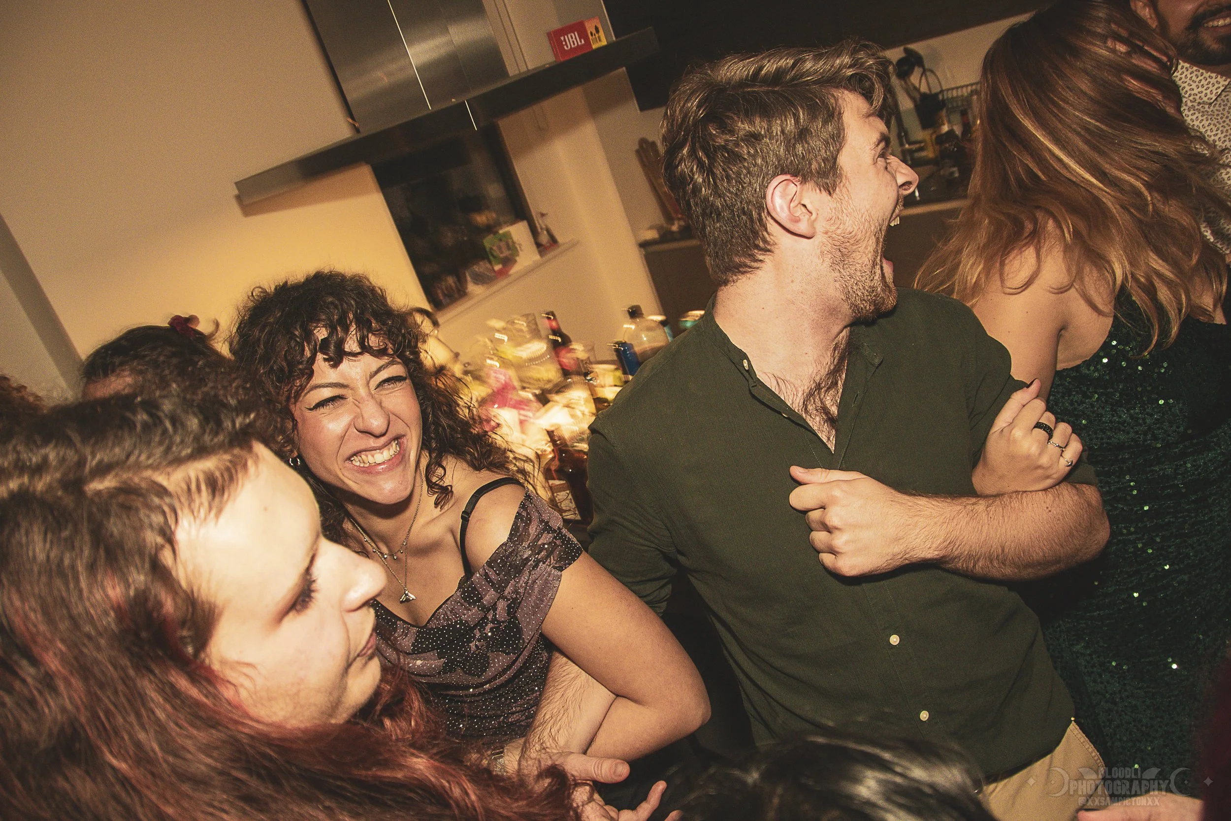 Group of people at a party, laughing and enjoying themselves in a kitchen.