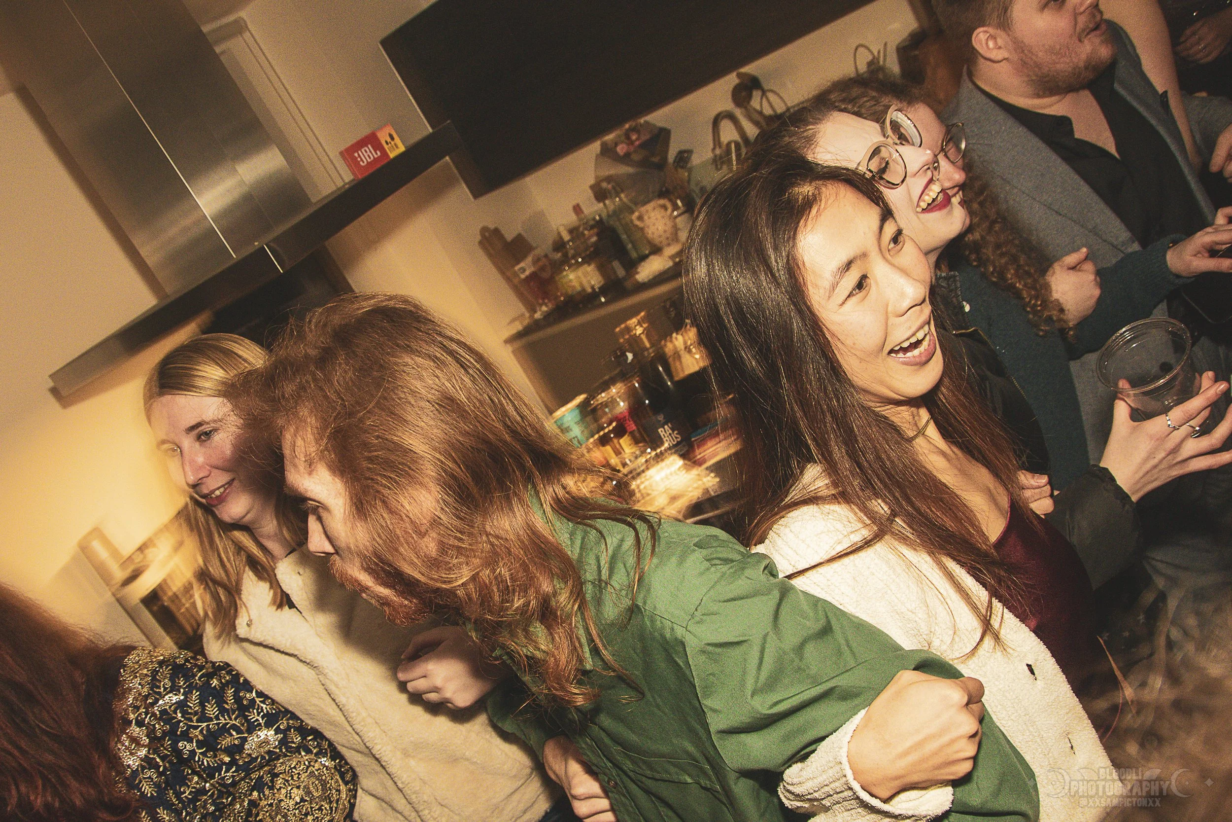 People enjoying a social gathering in a kitchen, smiling and holding drinks.
