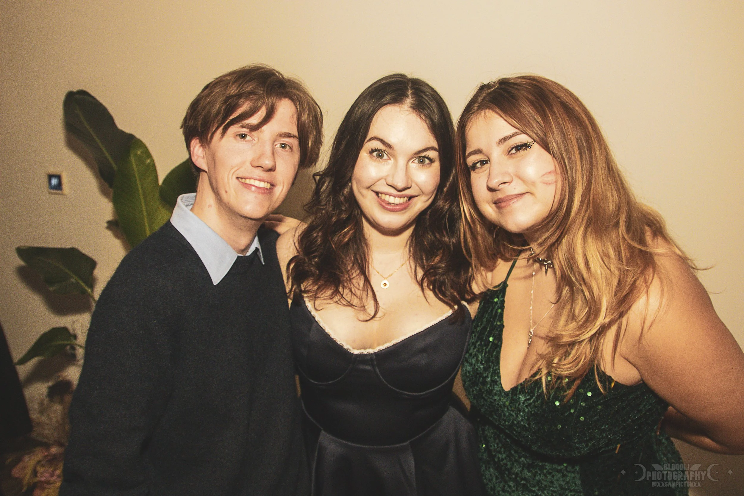 Three young adults smiling at a party, standing close together in front of a beige wall with large green plant leaves in the background.