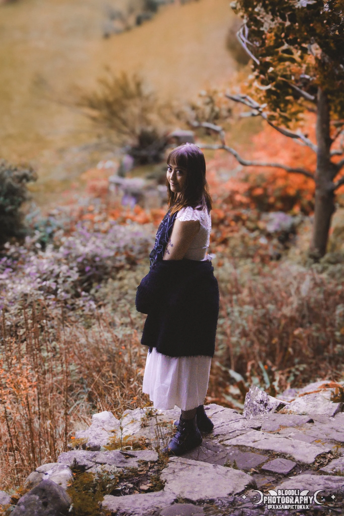 A woman standing on a rocky path in a forest with autumn foliage, wearing a white dress, black boots, and a black sweater draped over her shoulders, looking at the camera.