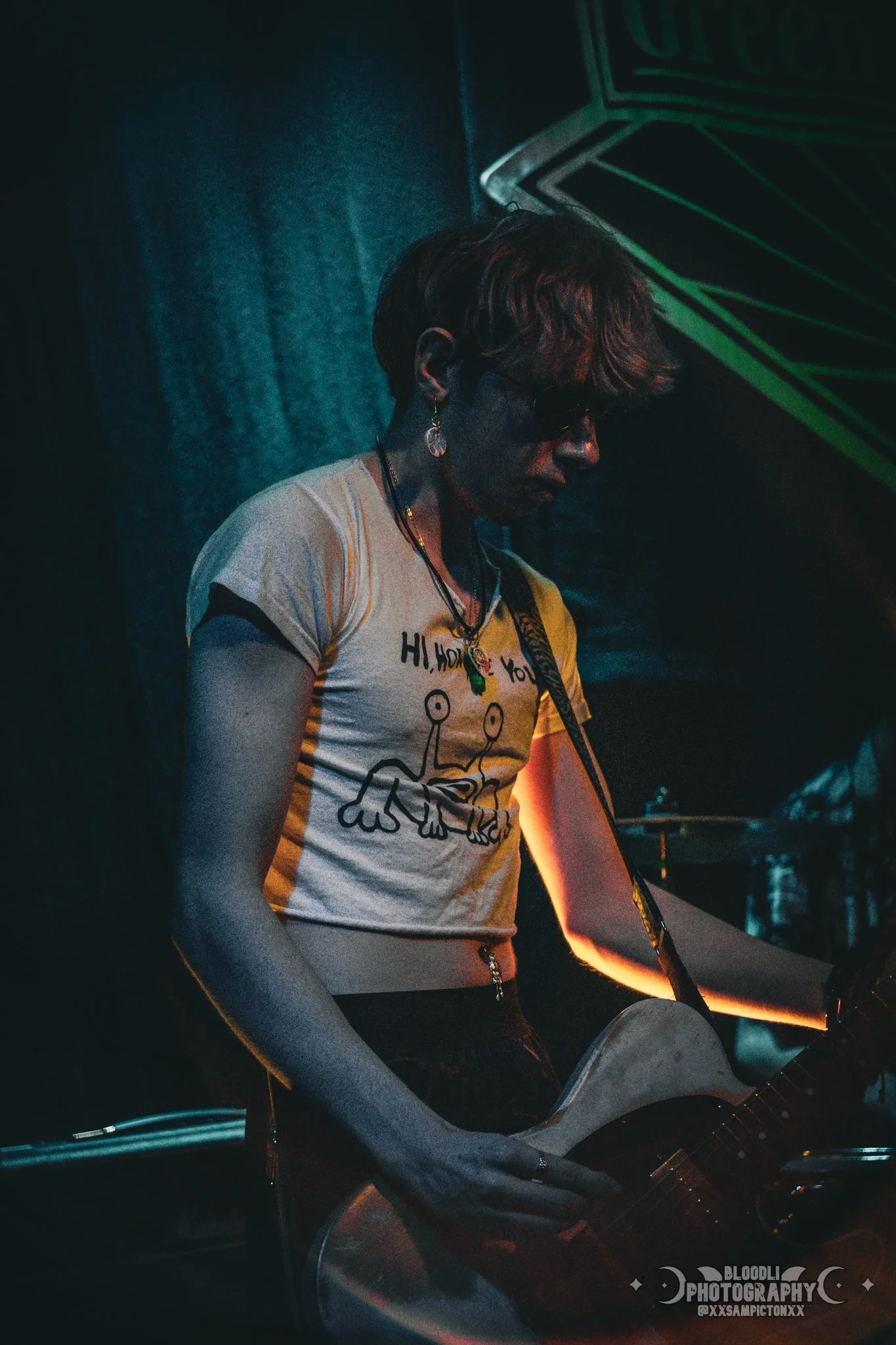 Person playing guitar on stage, wearing sunglasses and a T-shirt with a cartoon crab design in a dimly lit setting.